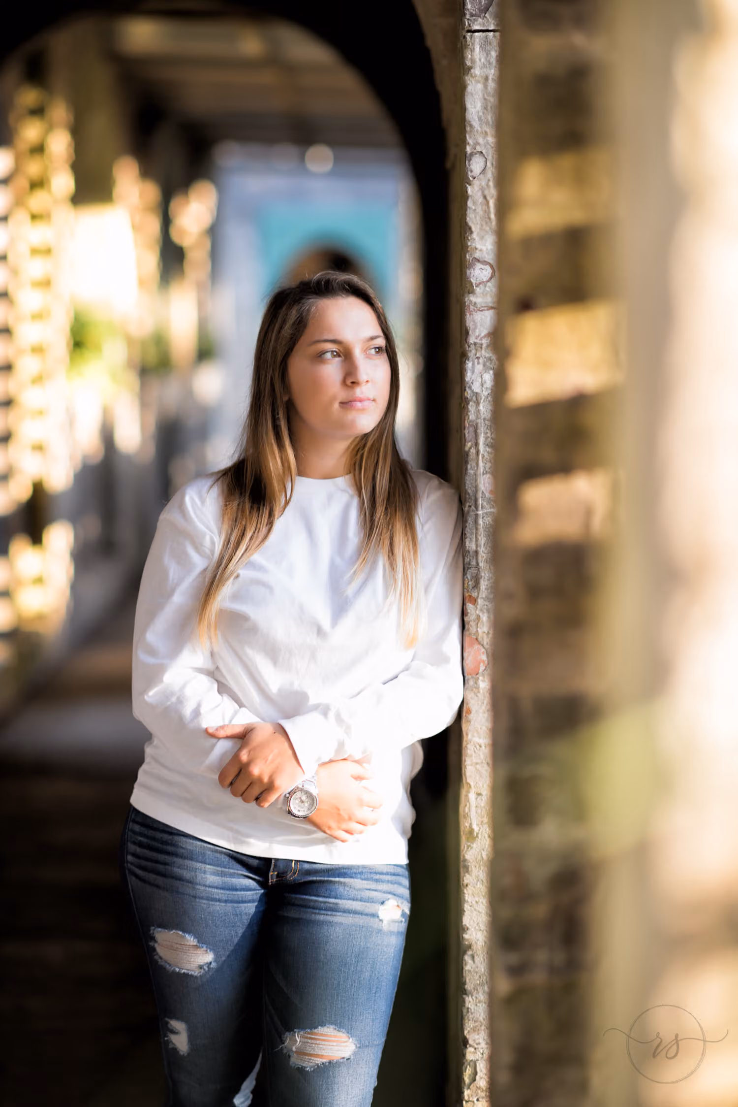 Senior in a white top and ripped jeans leaning on a pillar in the shaded arched hallway at Atalaya Castle