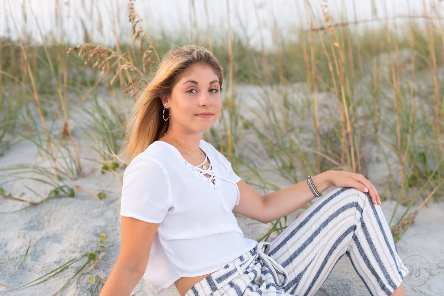 Senior in a white top and striped pants sitting by the dunes and sea oats at Huntington Beach State Park