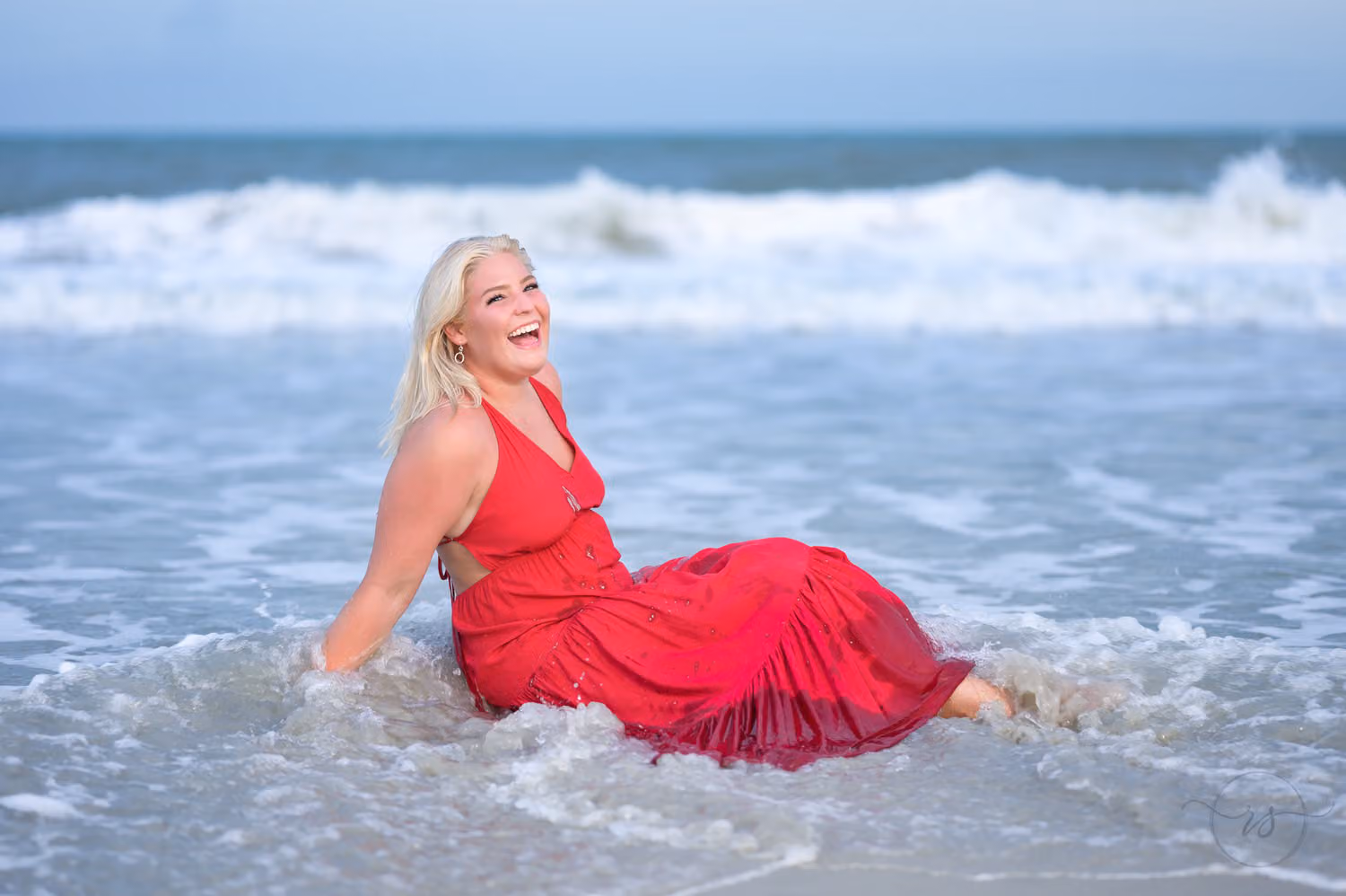 Senior in a red dress laughing as a wave washes over her where she sits in the surf at Huntington Beach State Park