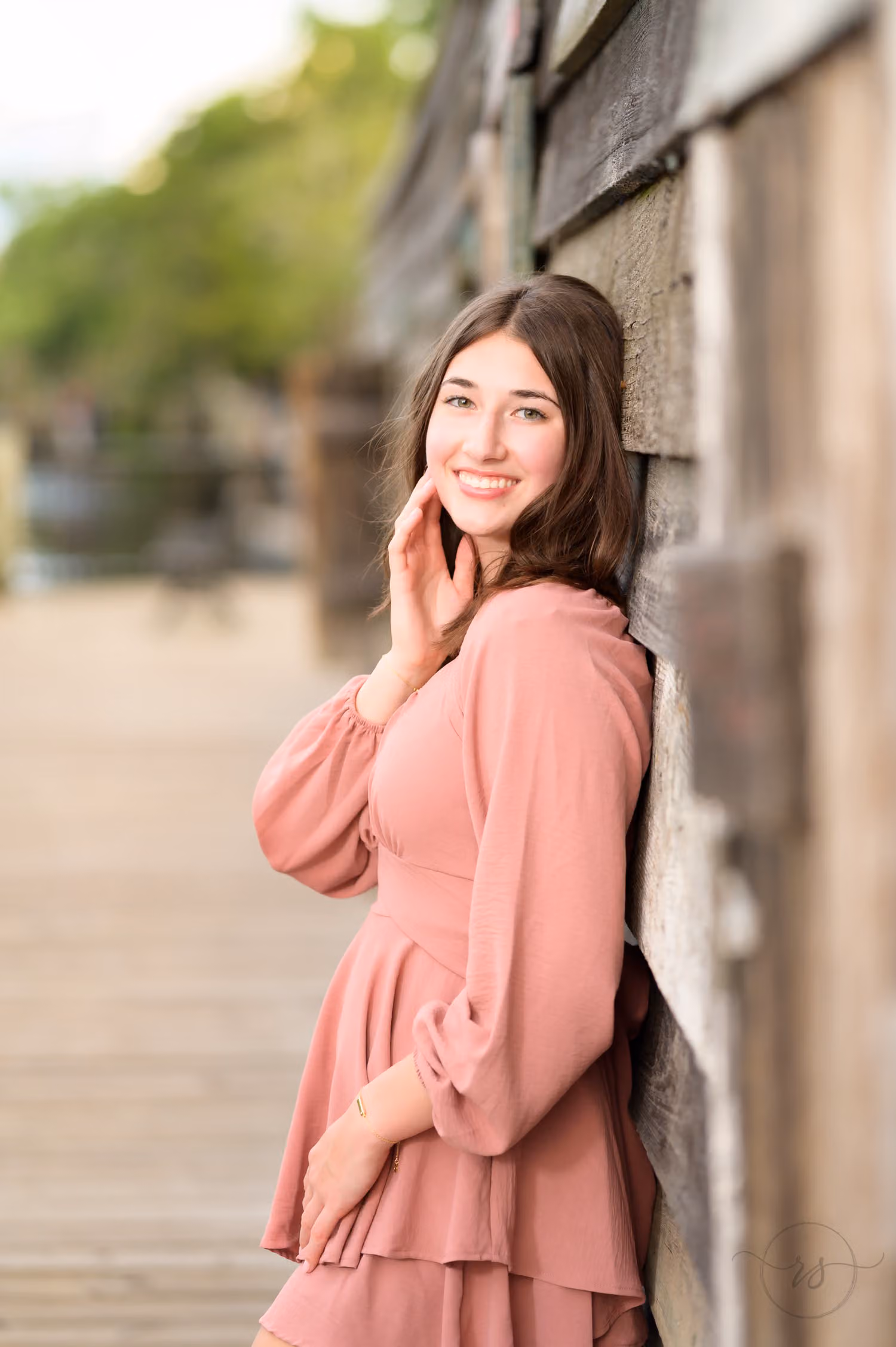 Senior girl in a pink dress leaning against a weathered wooden wall on the boardwalk at the Conway River Walk