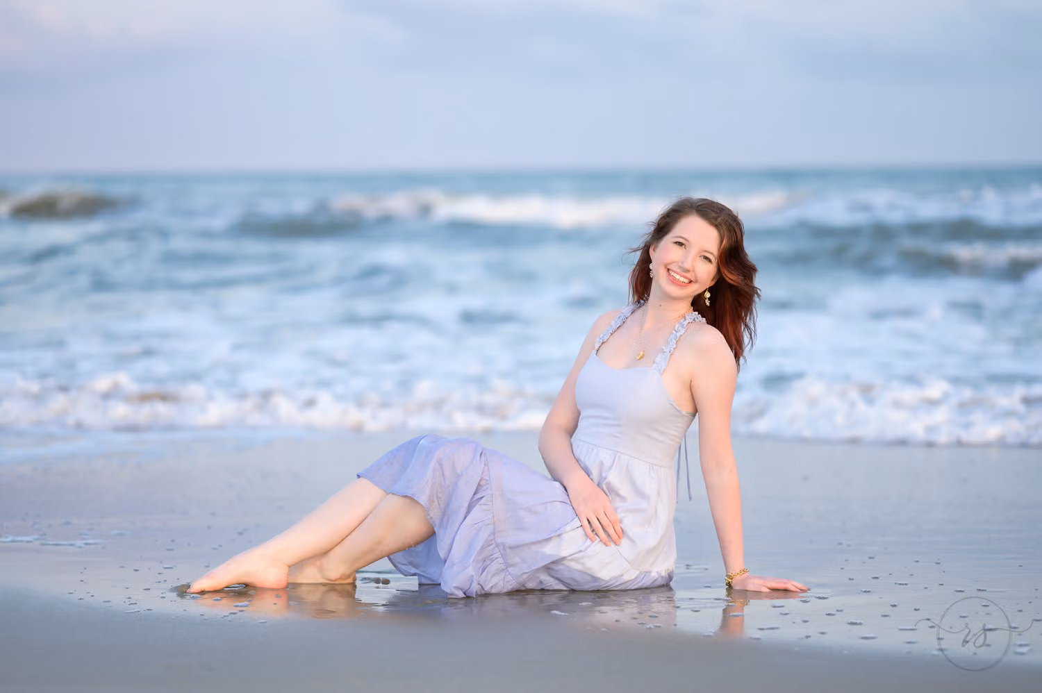 Senior girl in a light blue dress reclining in the shallow water as waves roll in behind her in Pawleys Island