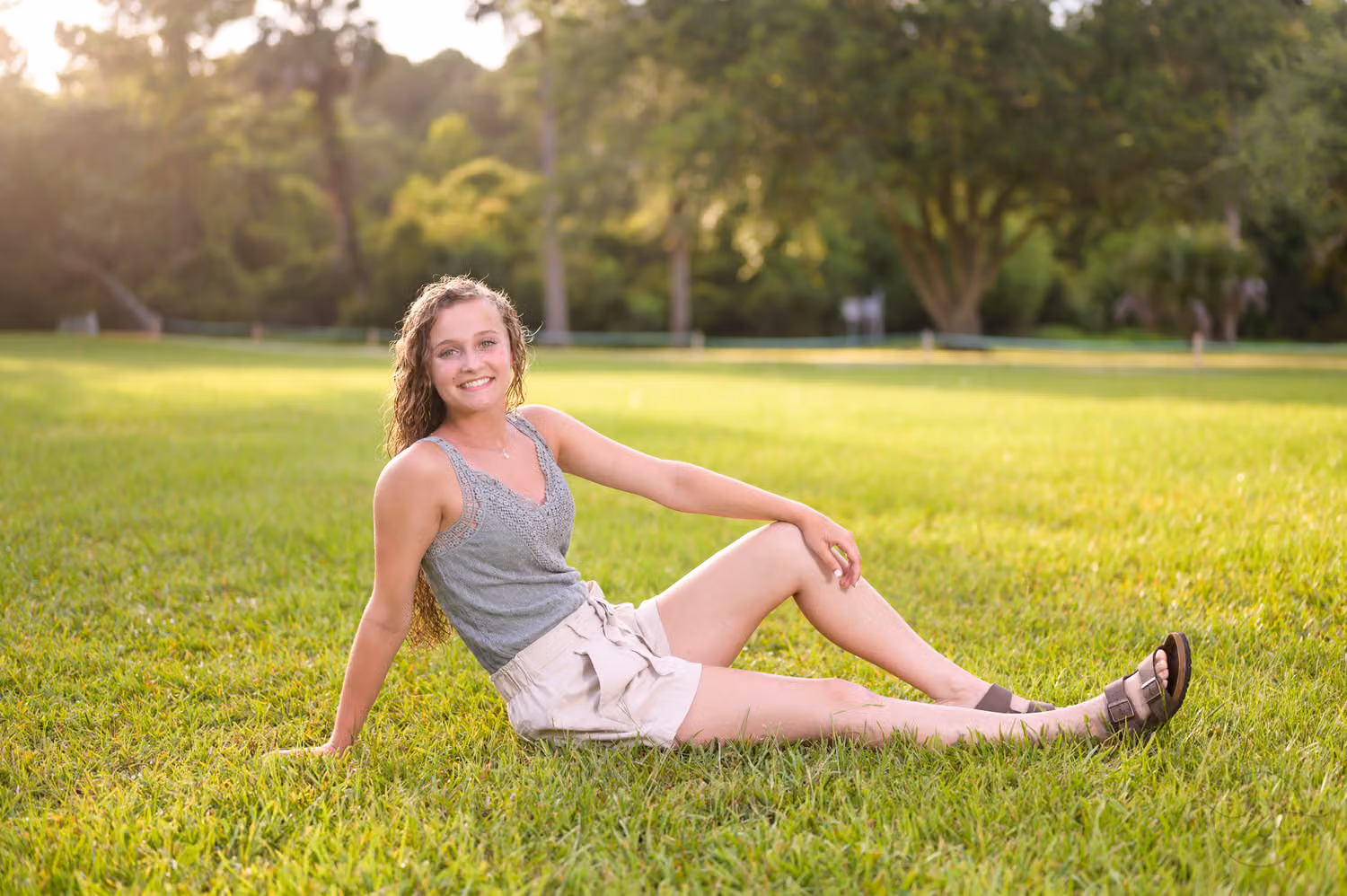 Senior girl in a gray tank top and shorts reclining on the green lawn under the trees at Huntington Beach State Park