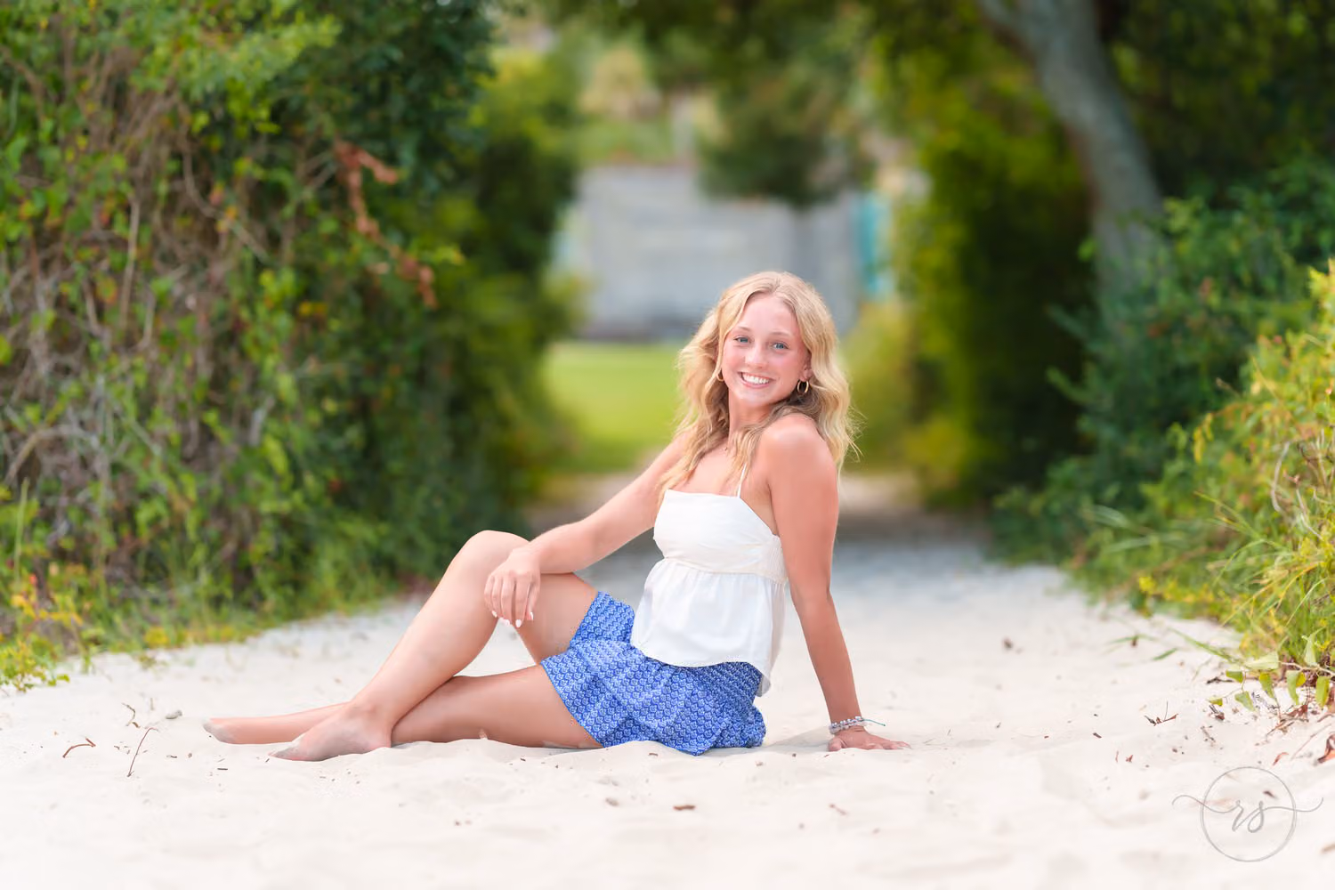 Senior girl in a white top and blue skirt reclining on the sandy path under the trees at Huntington Beach State Park