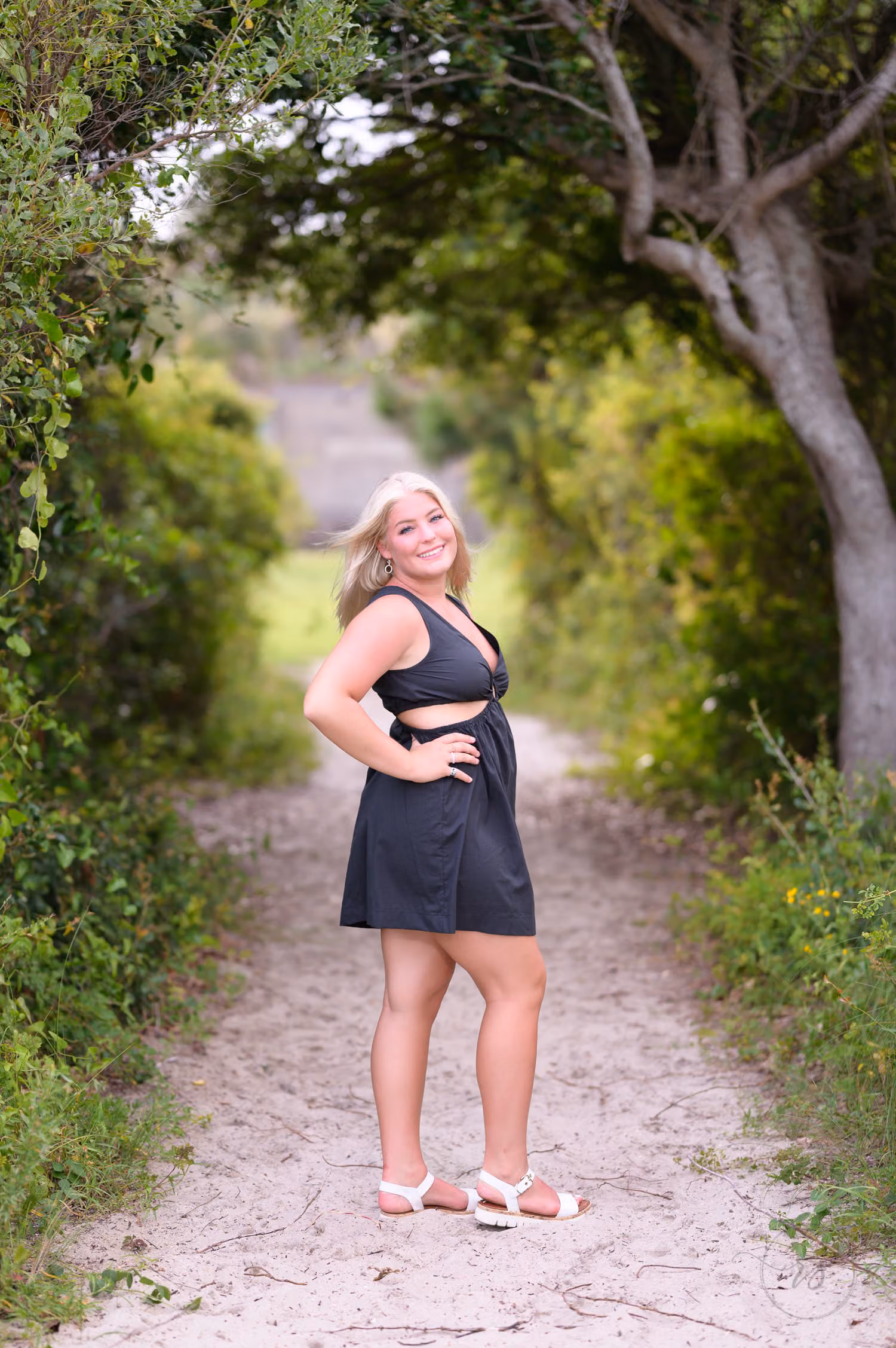 Senior in a black romper on a sandy path framed by an arch of trees at Huntington Beach State Park