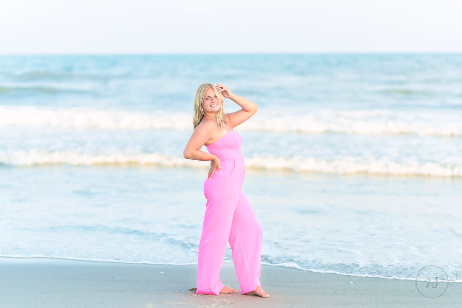 Senior girl in a pink jumpsuit posing with her hand in her hair in the shallow waves in North Myrtle Beach