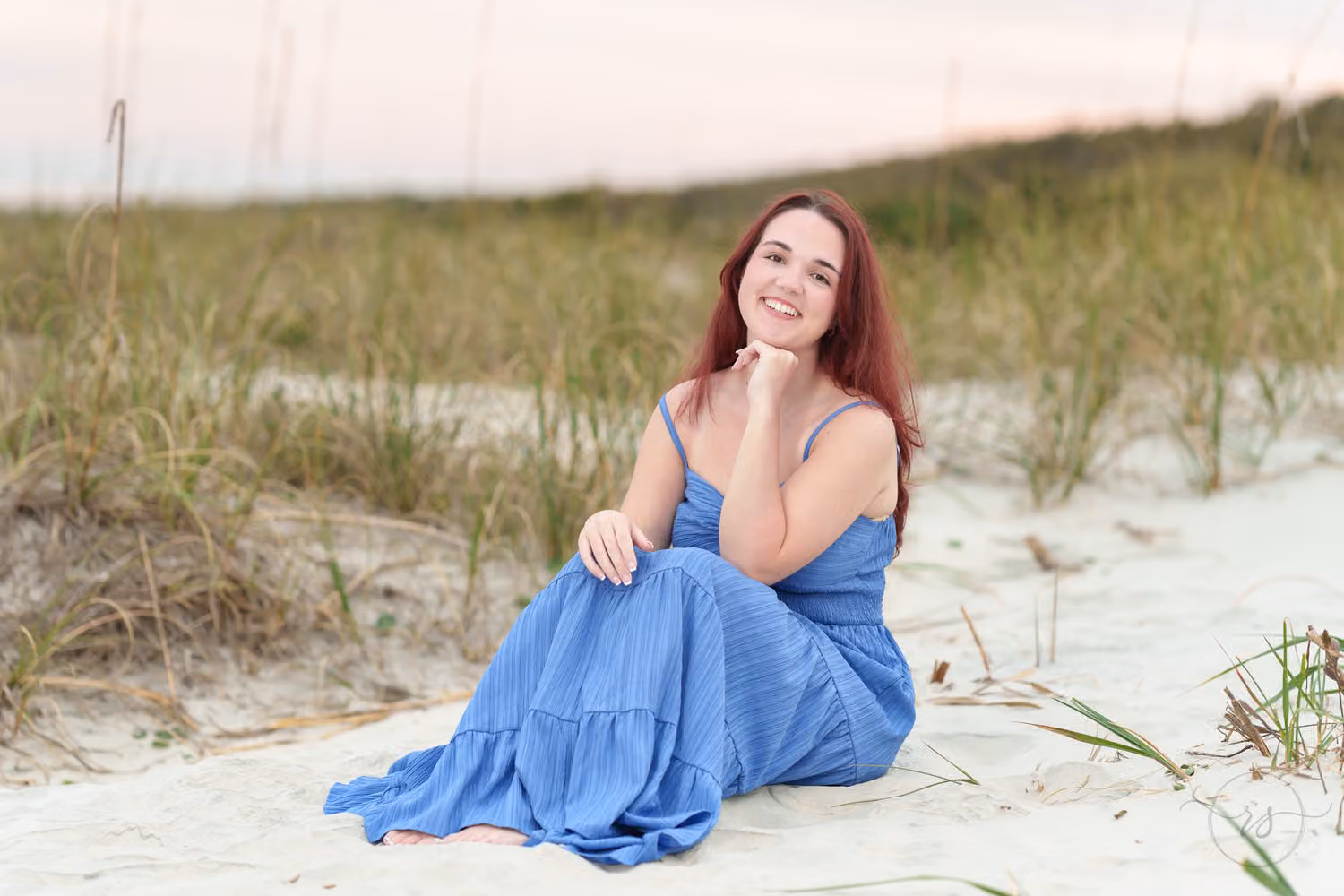 Senior in a blue dress sitting in the sand by the dune grass with her chin on her hand at Huntington Beach State Park