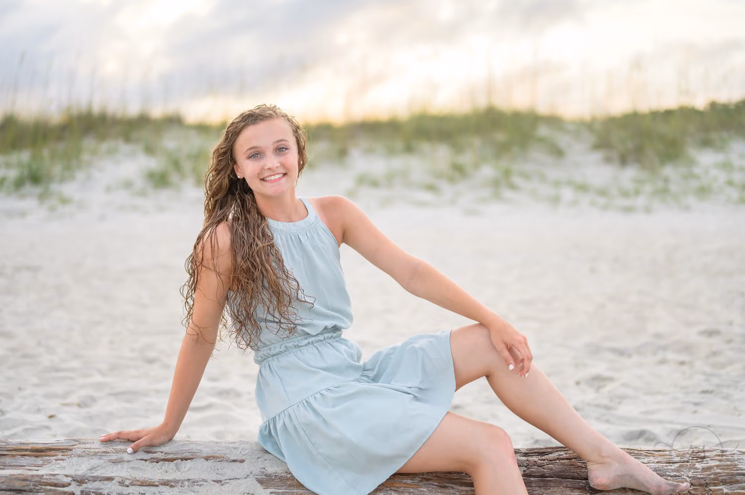 Senior girl in a mint dress sitting on driftwood by the dunes in soft sunset light at Huntington Beach State Park