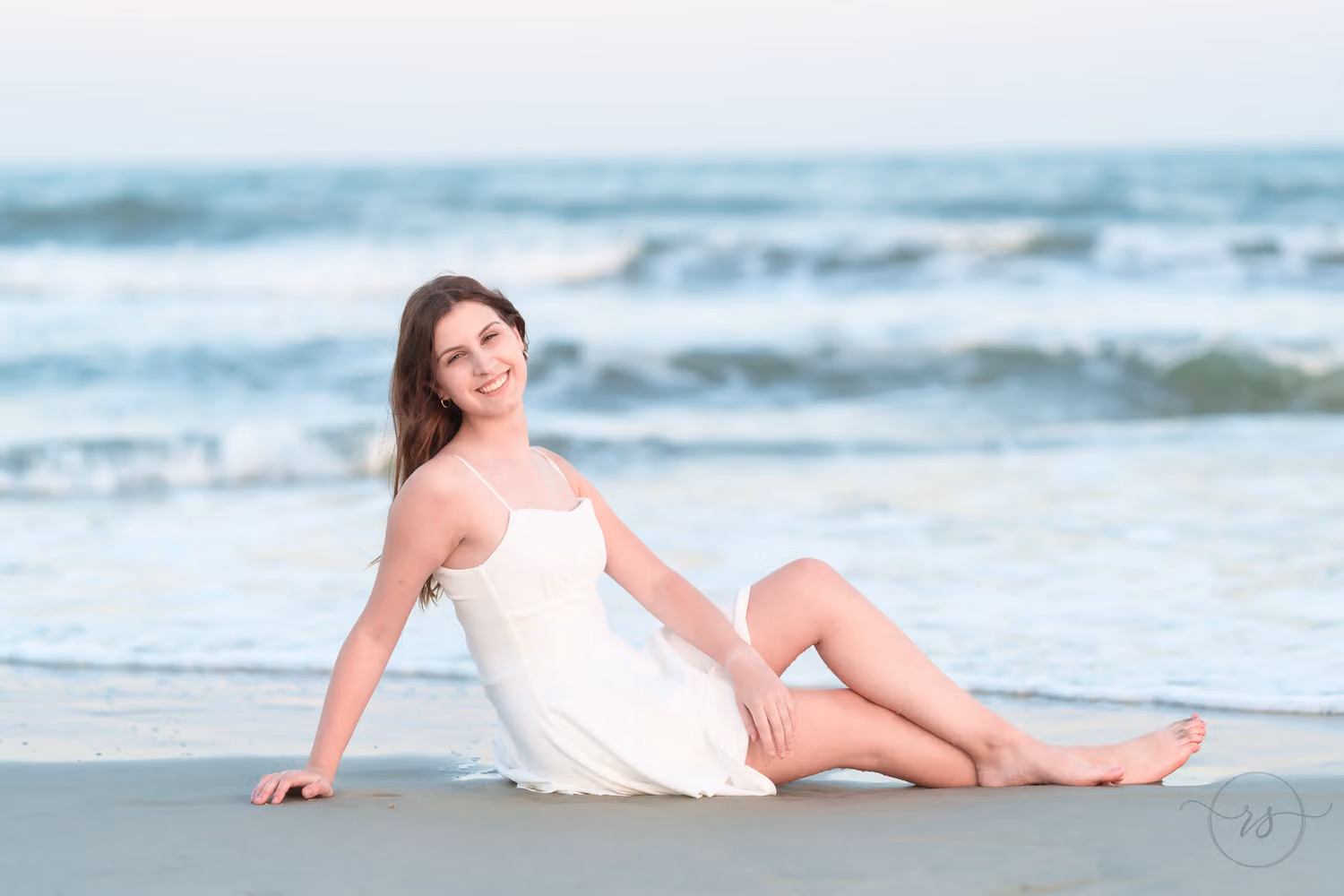 Senior girl in a white dress sitting in the sand by the waves at Huntington Beach State Park