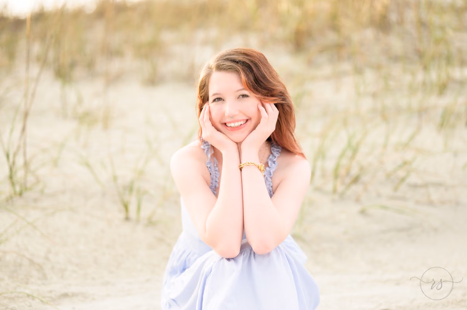 Smiling senior girl sitting in the sand by the dune grass with her hands framing her face in Pawleys Island