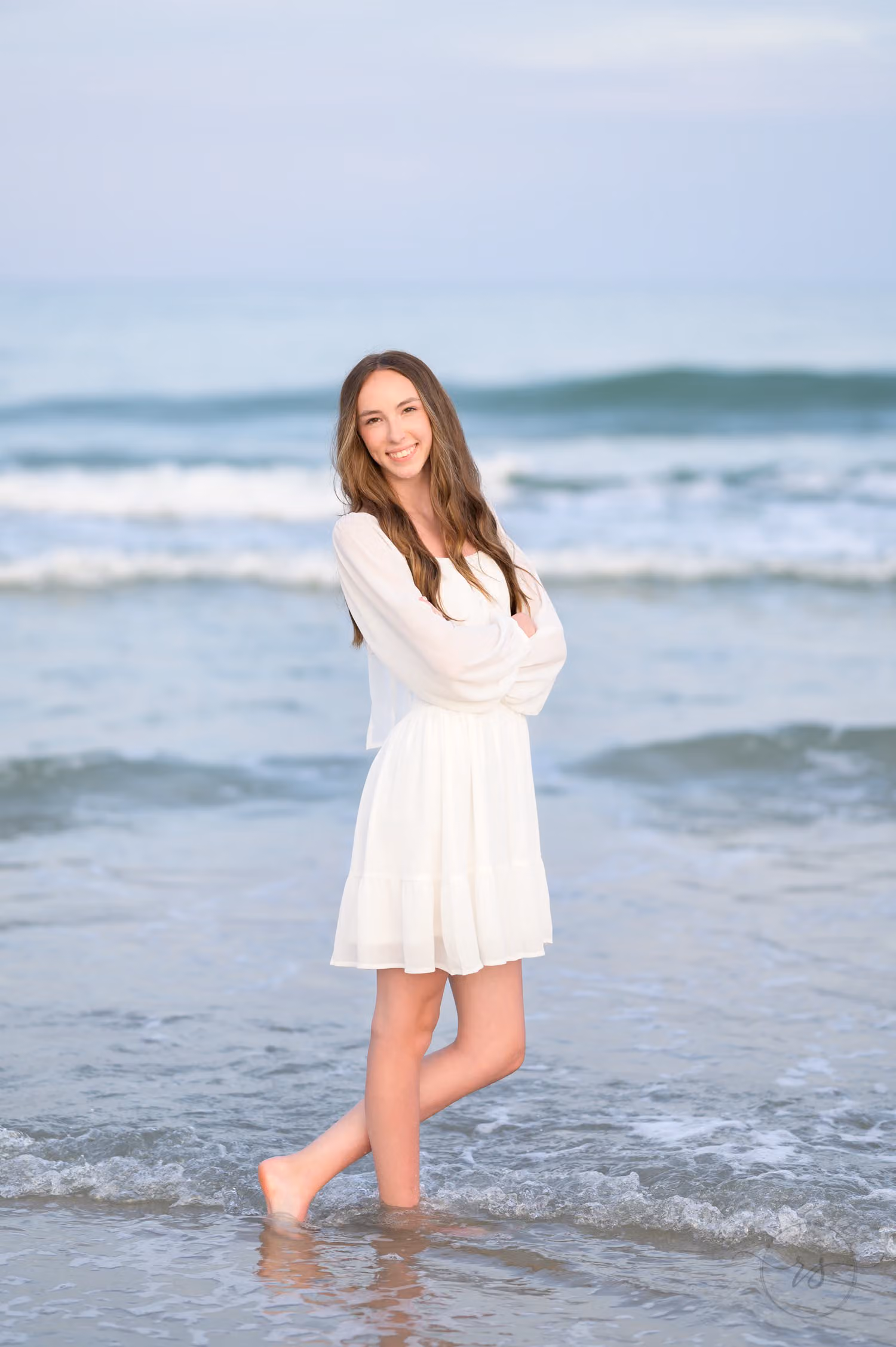 High school senior in a white dress standing barefoot in the shallow surf at Huntington Beach State Park
