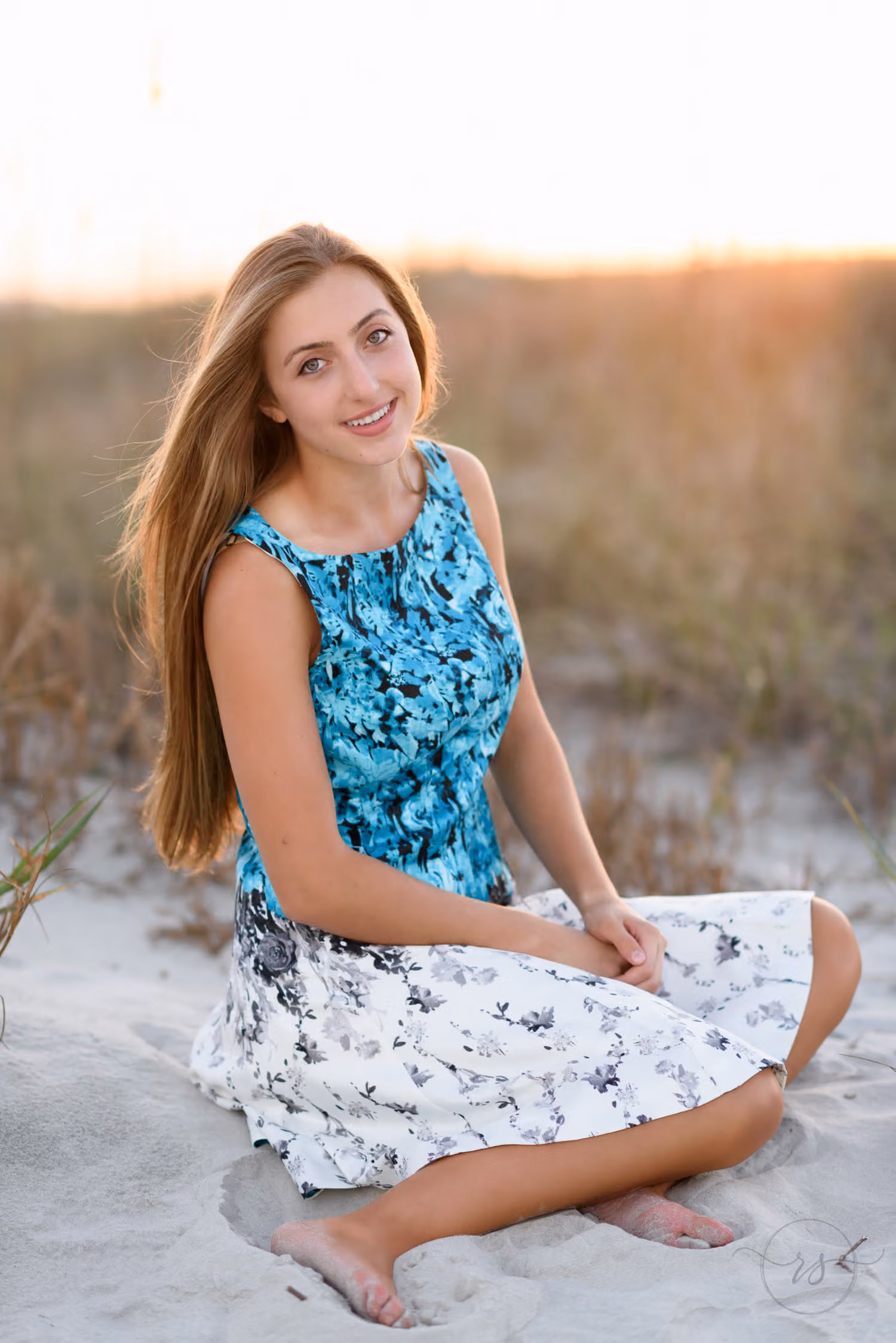 Senior in a teal and white floral dress sitting cross-legged in the sand at sunset at Huntington Beach State Park