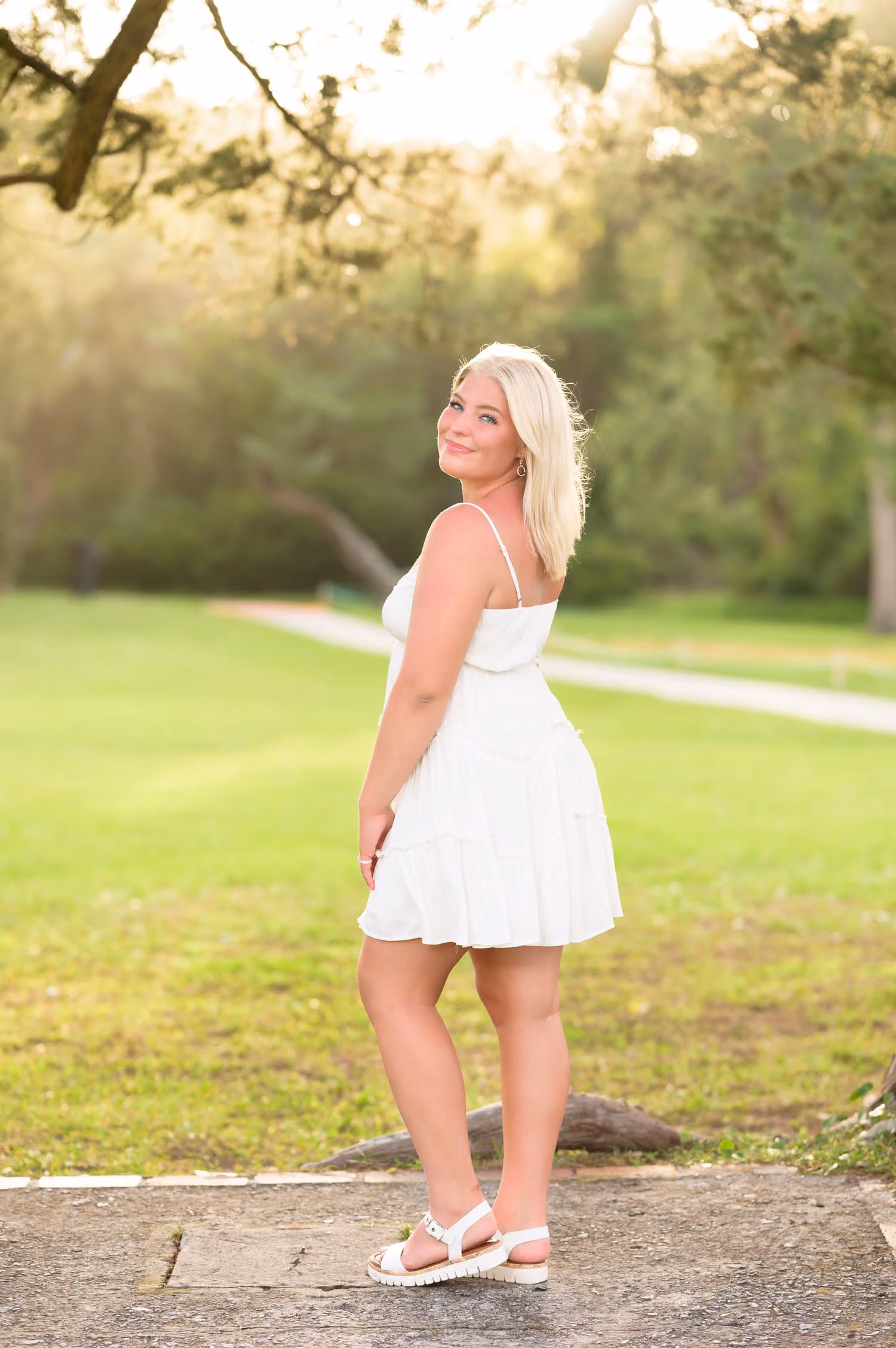 Blonde senior in a white sundress glowing in sunset backlight on a grassy lawn at Huntington Beach State Park