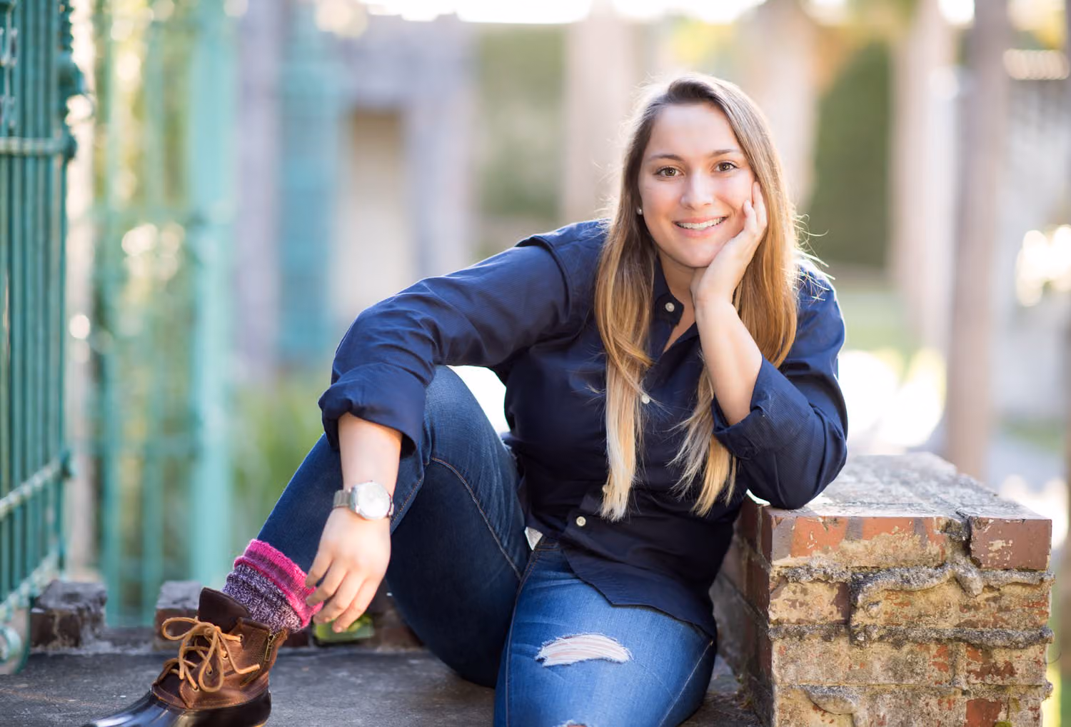 Senior in a navy shirt and duck boots sitting on a brick ledge with her chin on her hand at Atalaya Castle