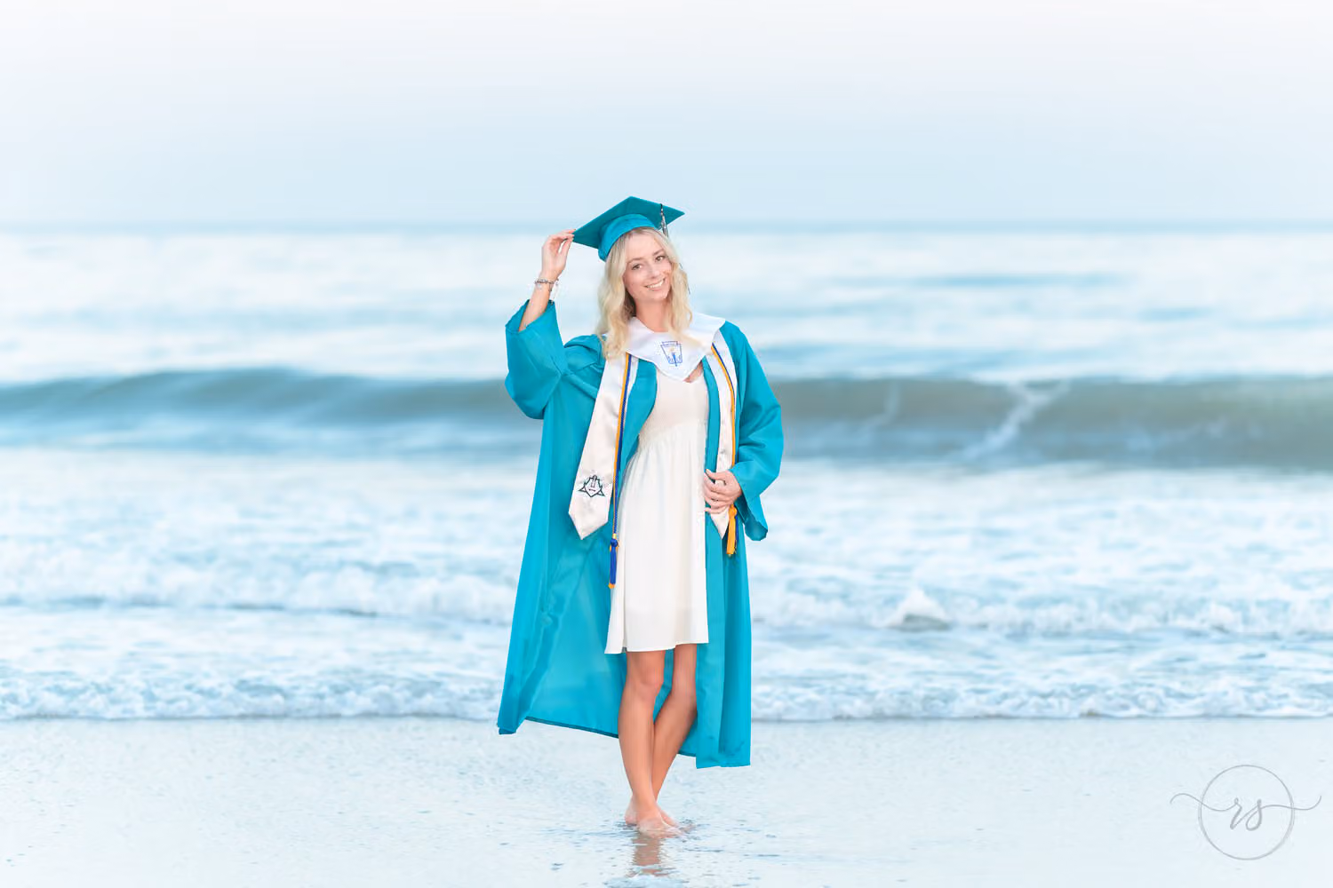 Senior in a teal graduation gown and cap standing at the water's edge with waves behind her at Huntington Beach State Park
