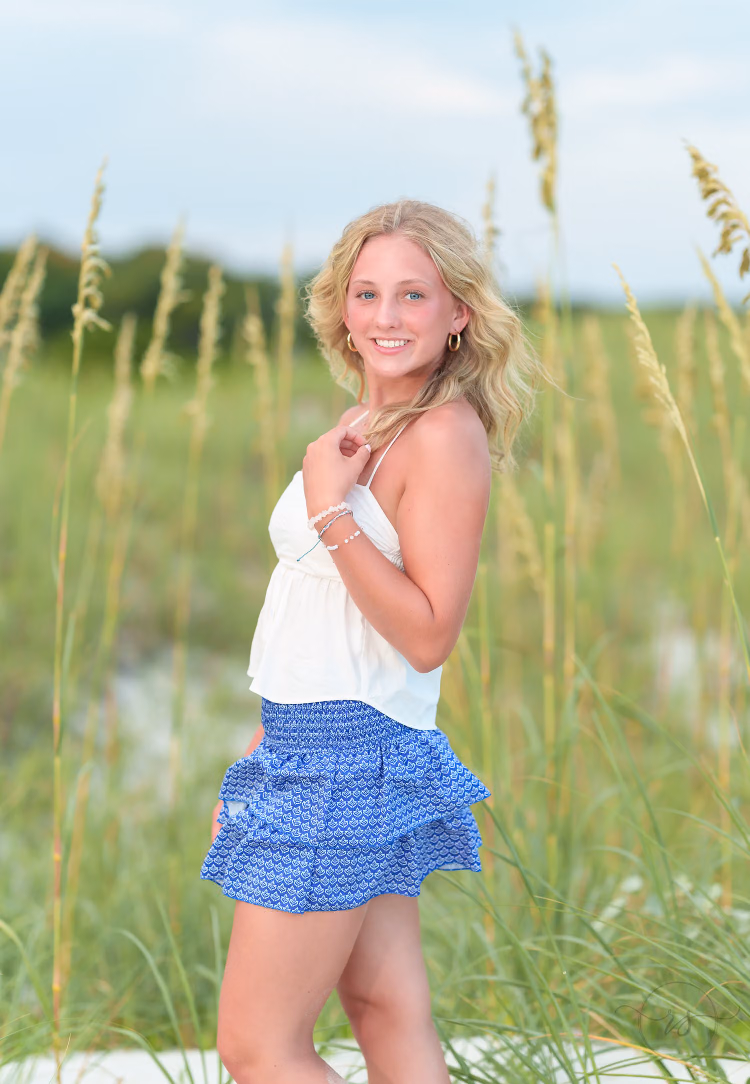 Senior girl in a white top and ruffled blue skirt smiling in front of tall sea oats at Huntington Beach State Park