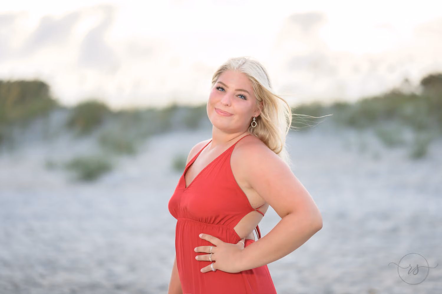Blonde senior in a red dress with her hand on her hip in front of the dunes at Huntington Beach State Park