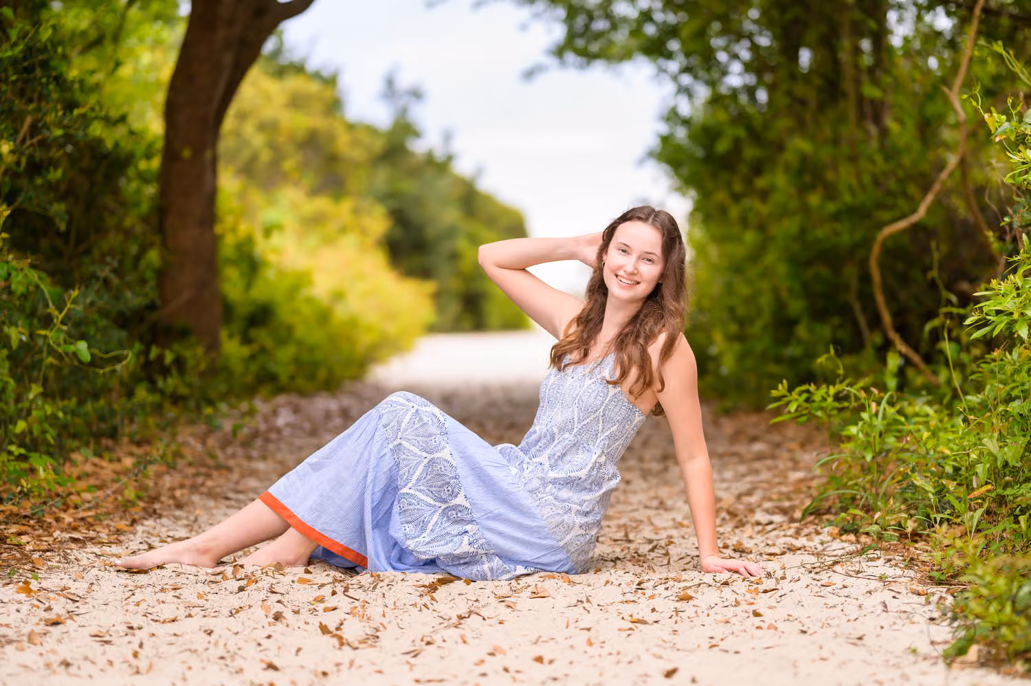 Senior in a lavender dress reclining on the sandy path under an arch of trees at Huntington Beach State Park