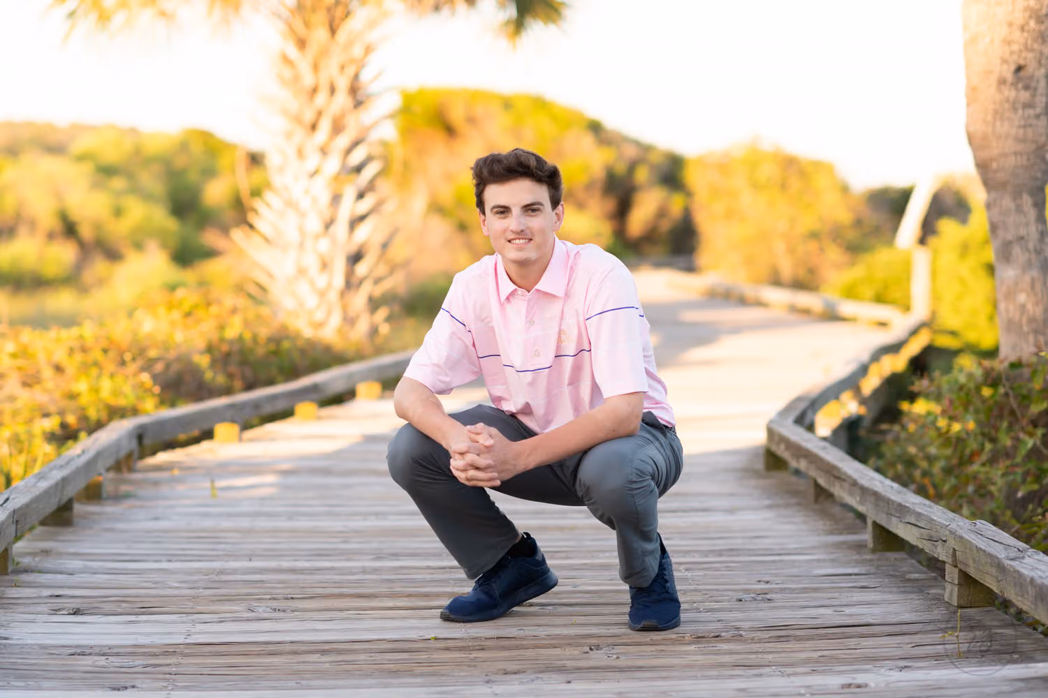 Senior guy in a pink shirt crouching on a beach boardwalk lined with palmettos at Myrtle Beach State Park