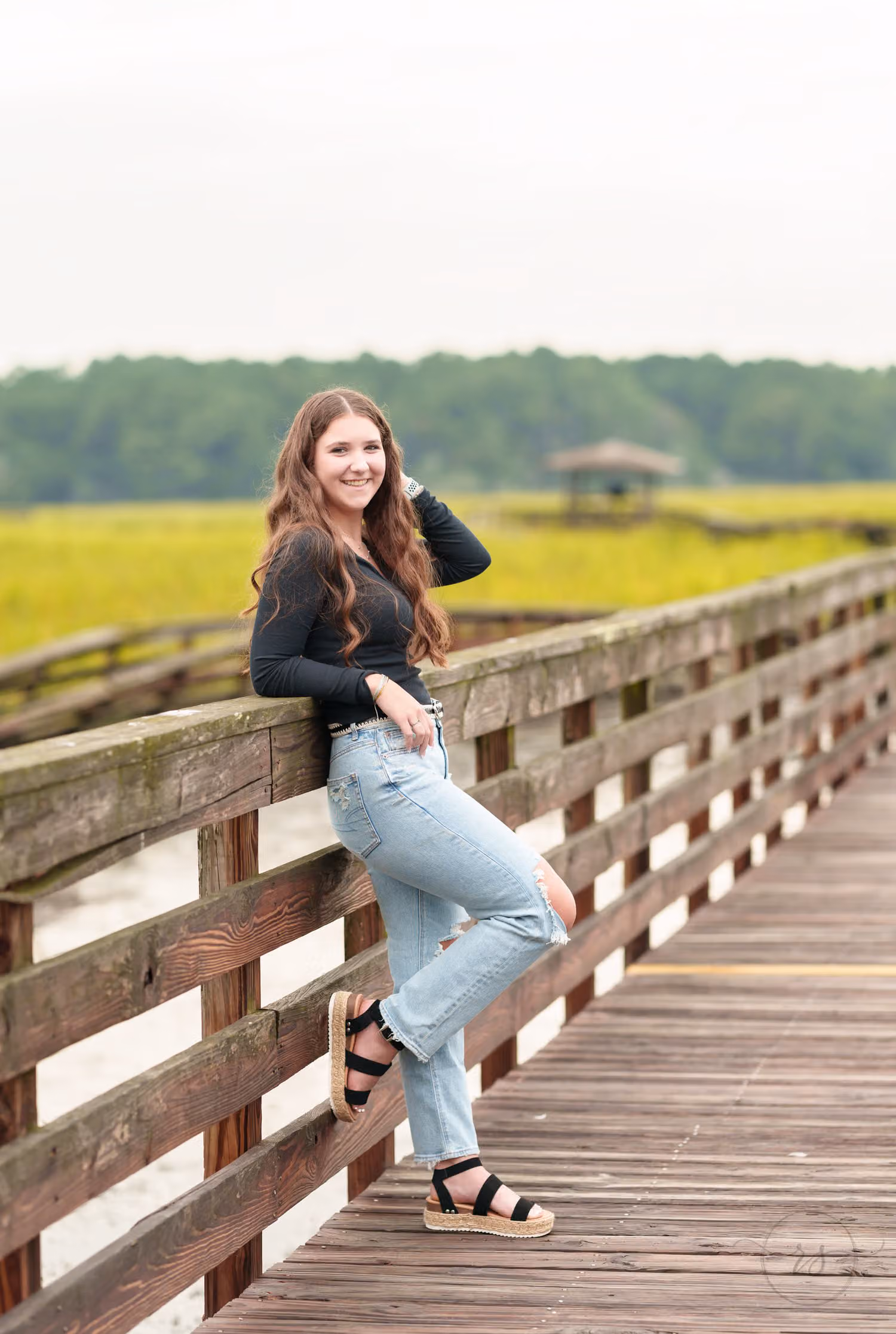 Senior with a hand in her hair resting against the boardwalk rail, green marsh behind at Huntington Beach State Park