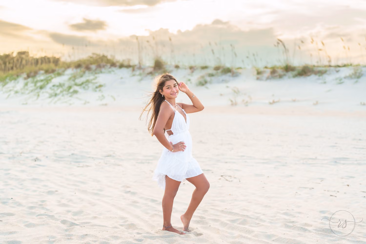 Senior in a white dress with her hand in her hair on the sand in front of the dunes at Huntington Beach State Park