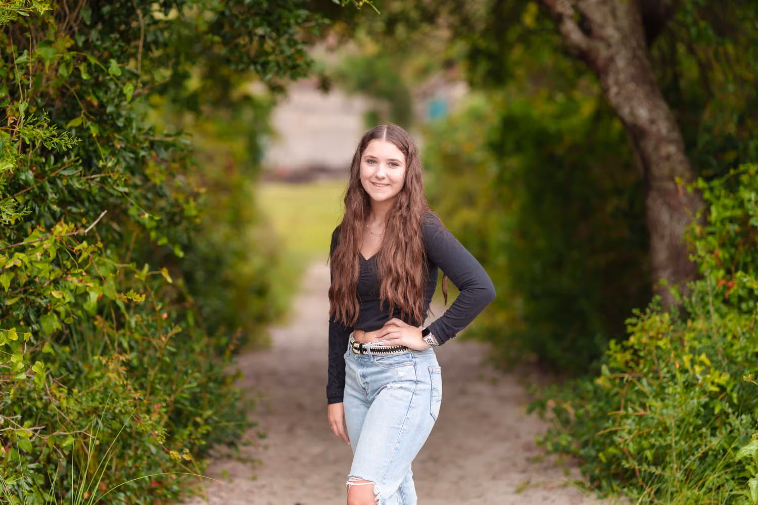 Senior in a black top and jeans with a hand on her hip on a shaded sandy path at Huntington Beach State Park