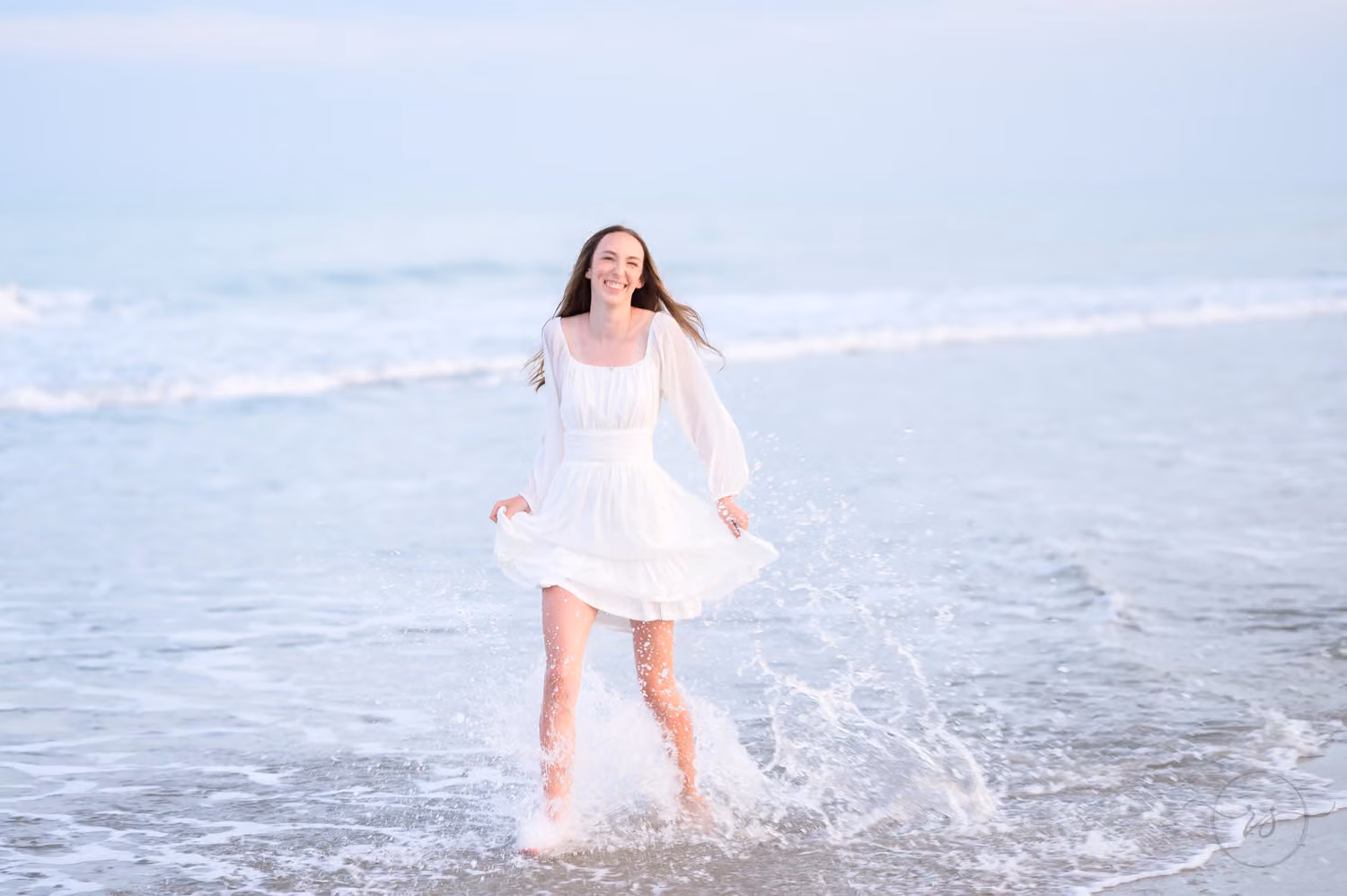 Laughing senior in a white dress kicking up splashes as she runs through the shallow surf at Huntington Beach State Park