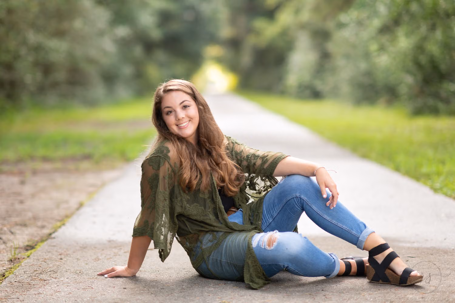 Senior in ripped jeans and a lace kimono sitting on a paved path lined with trees at Huntington Beach State Park