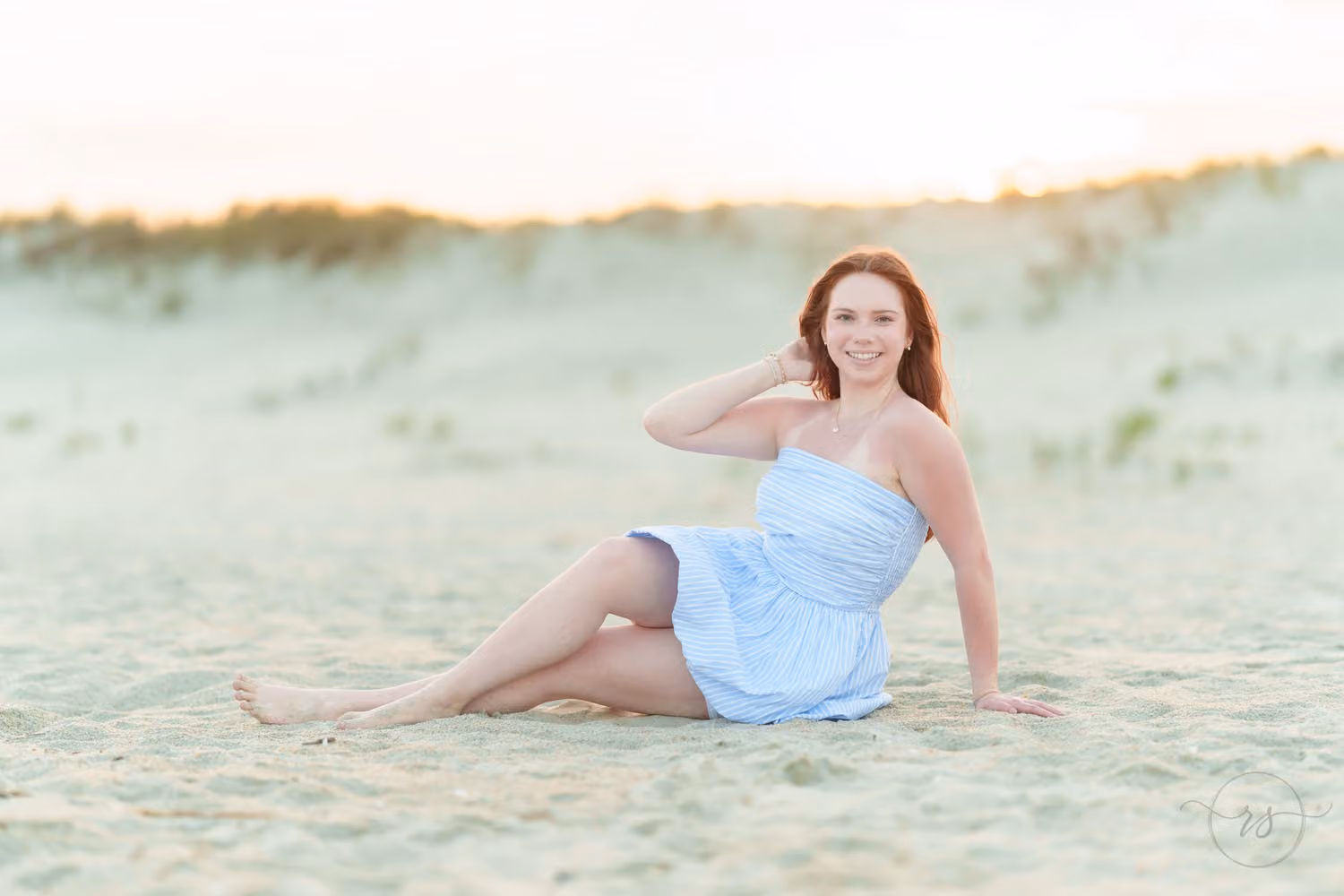 Senior girl in a light blue dress sitting in the sand with a hand in her hair by the dunes at Huntington Beach State Park