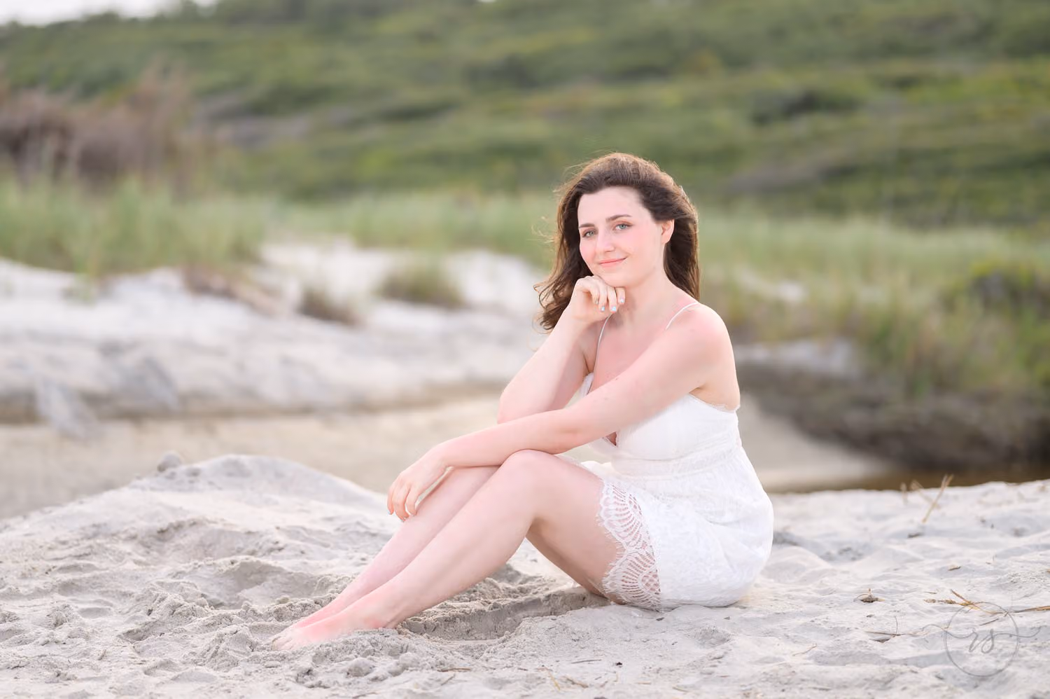 Senior in a white dress sitting in the sand hugging her knees beside the dunes at Myrtle Beach State Park