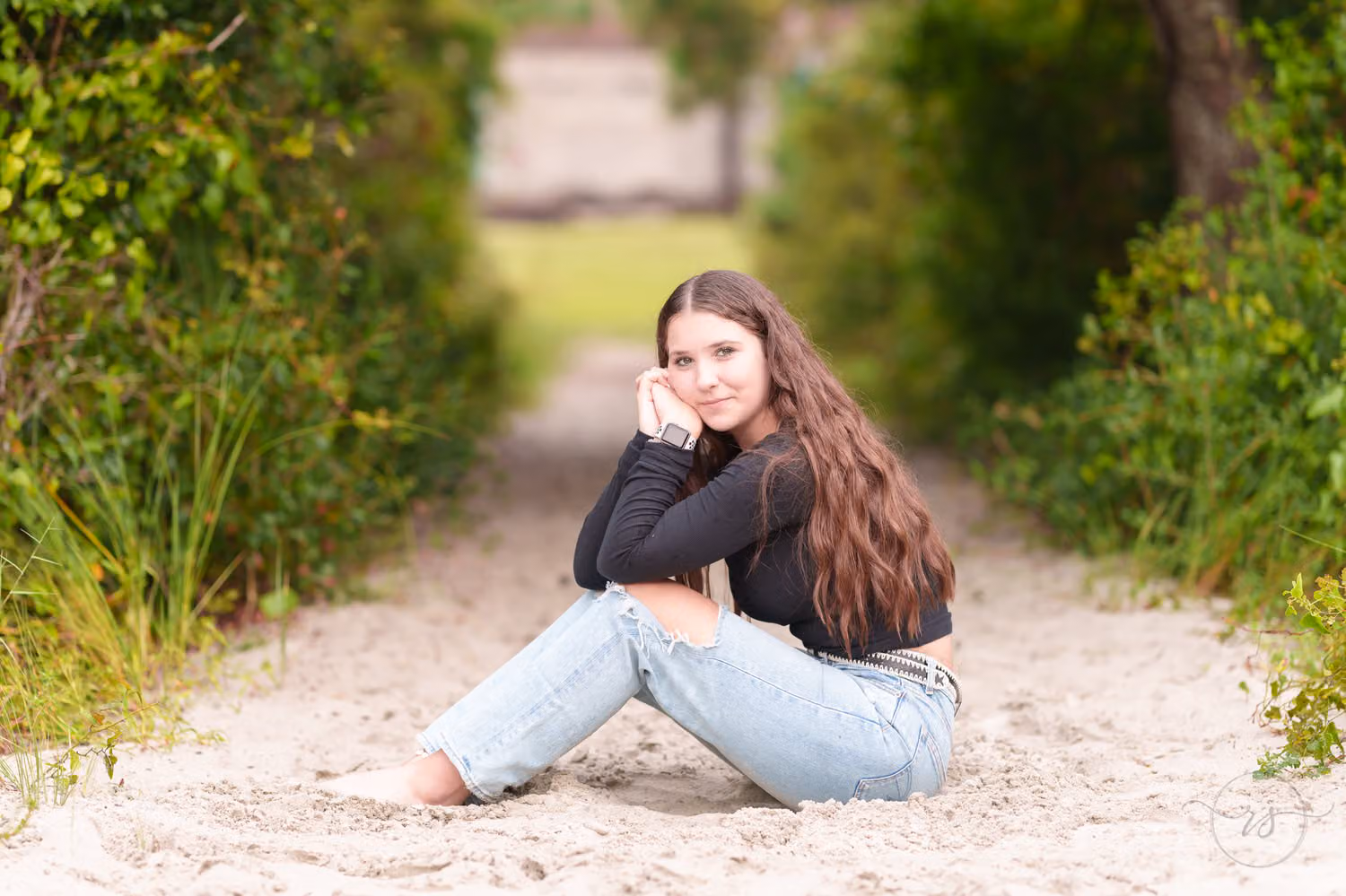 Senior in jeans sitting on the sandy trail with her cheek resting on her hand at Huntington Beach State Park