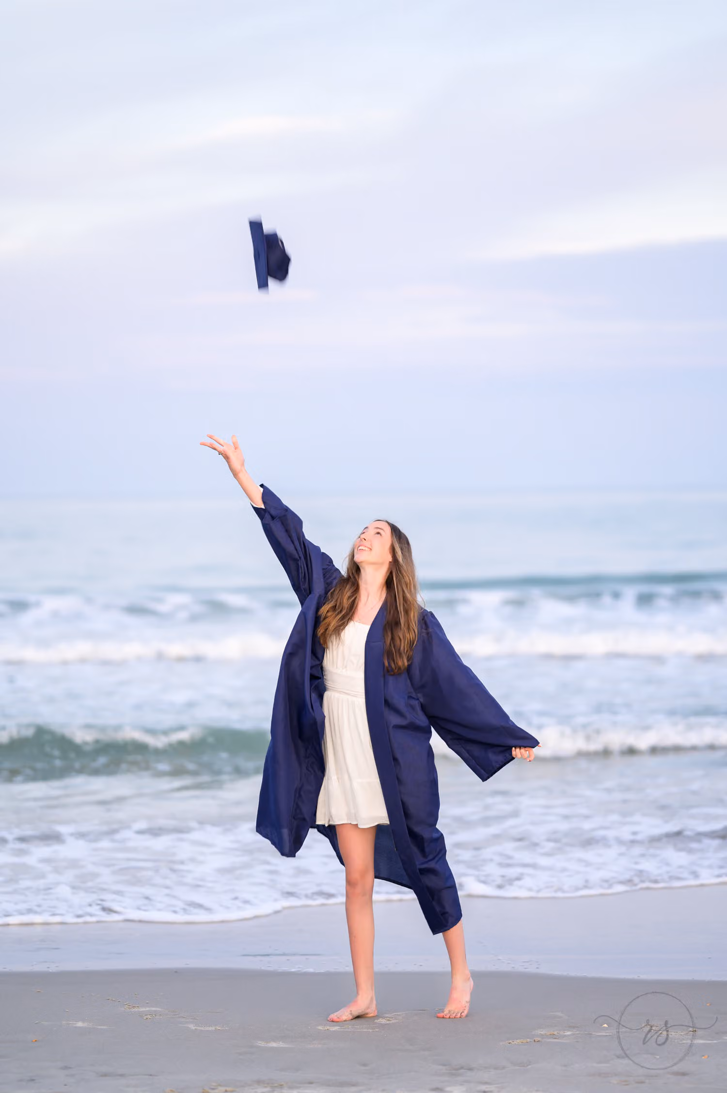 Graduate in a navy gown tossing her cap high over the beach at dusk at Huntington Beach State Park