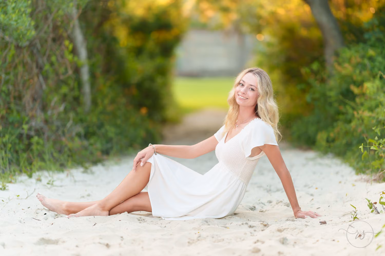 Senior girl in a white dress sitting in the sand on the tree-lined path to the beach at Huntington Beach State Park