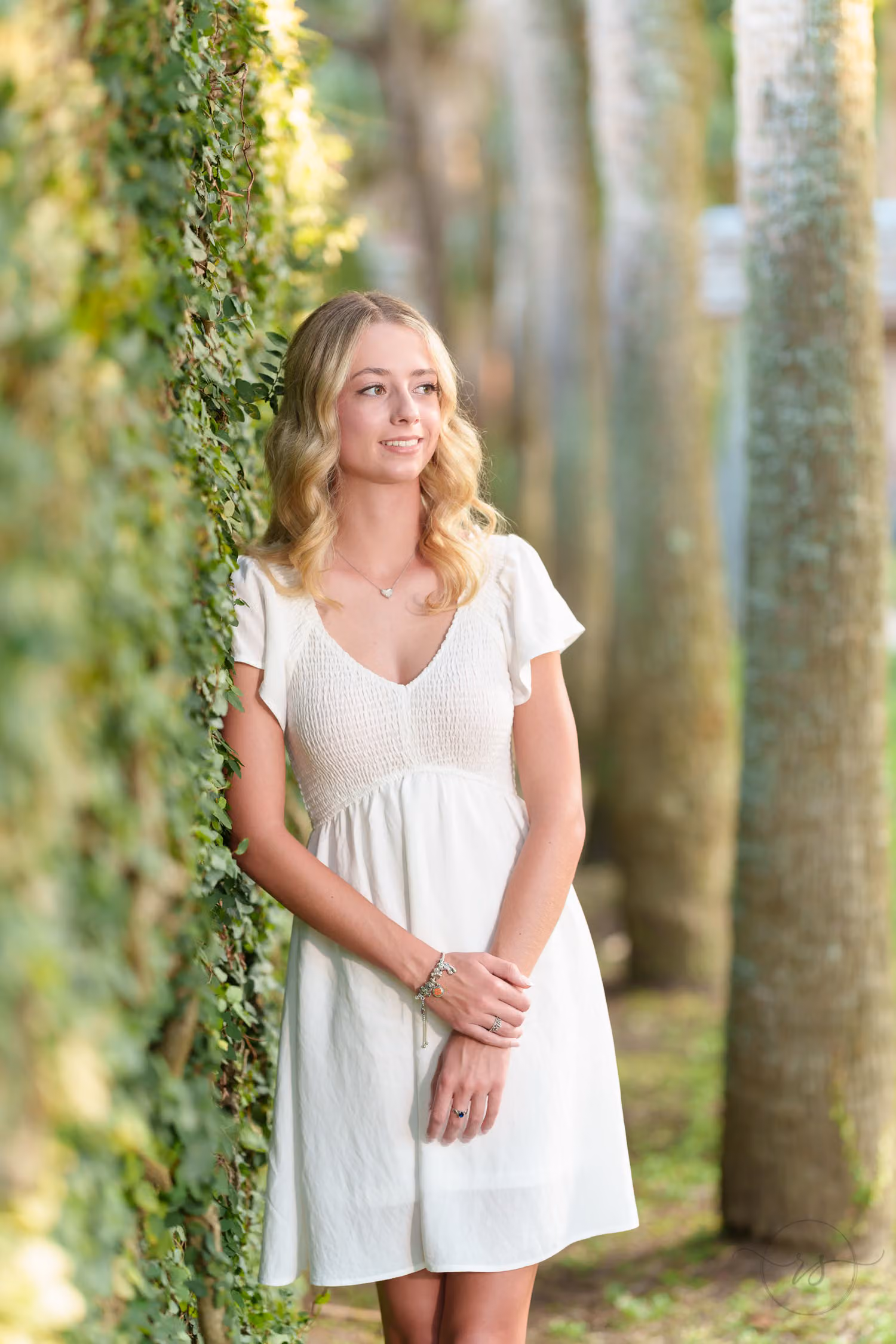 Senior girl in a white dress leaning against the ivy-covered wall among the palm trunks at Huntington Beach State Park