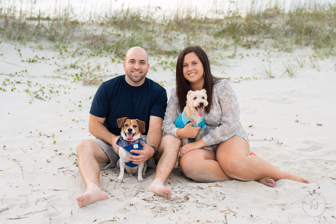 Couple sitting in the sand by the dunes with their two dogs at Huntington Beach State Park