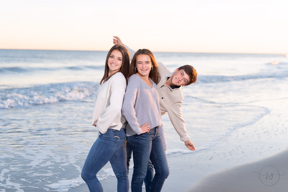 Three adult kids strike playful poses together at the water's edge at Huntington Beach State Park