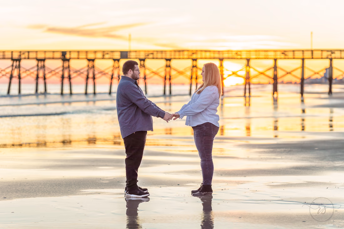 Couple holding hands on the wet sand in front of a fishing pier at sunset