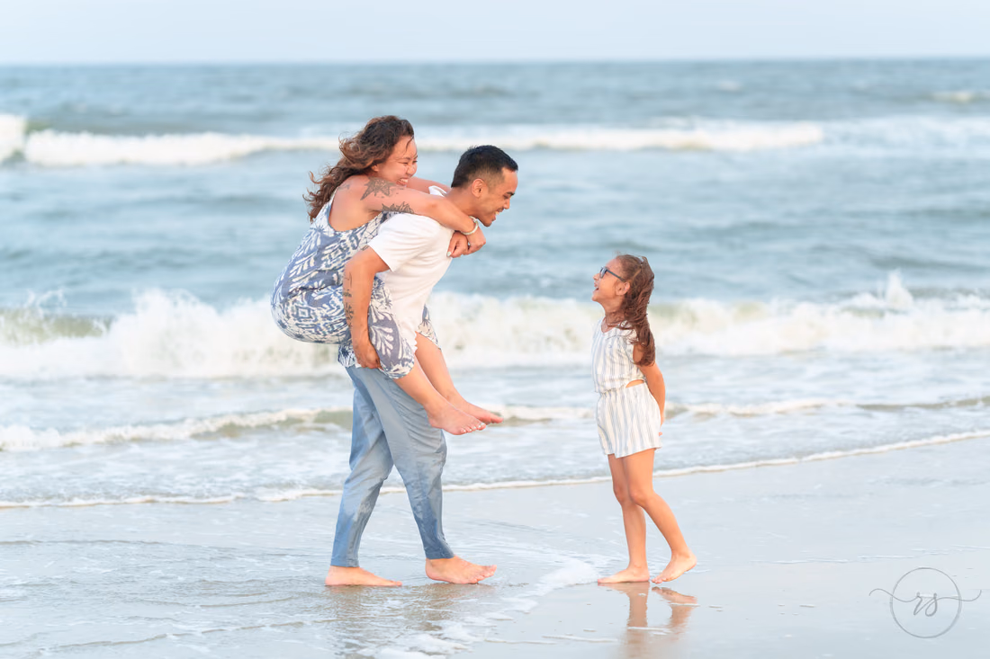 Daughter laughing as dad gives mom a piggyback ride in the surf - Huntington Beach State Park