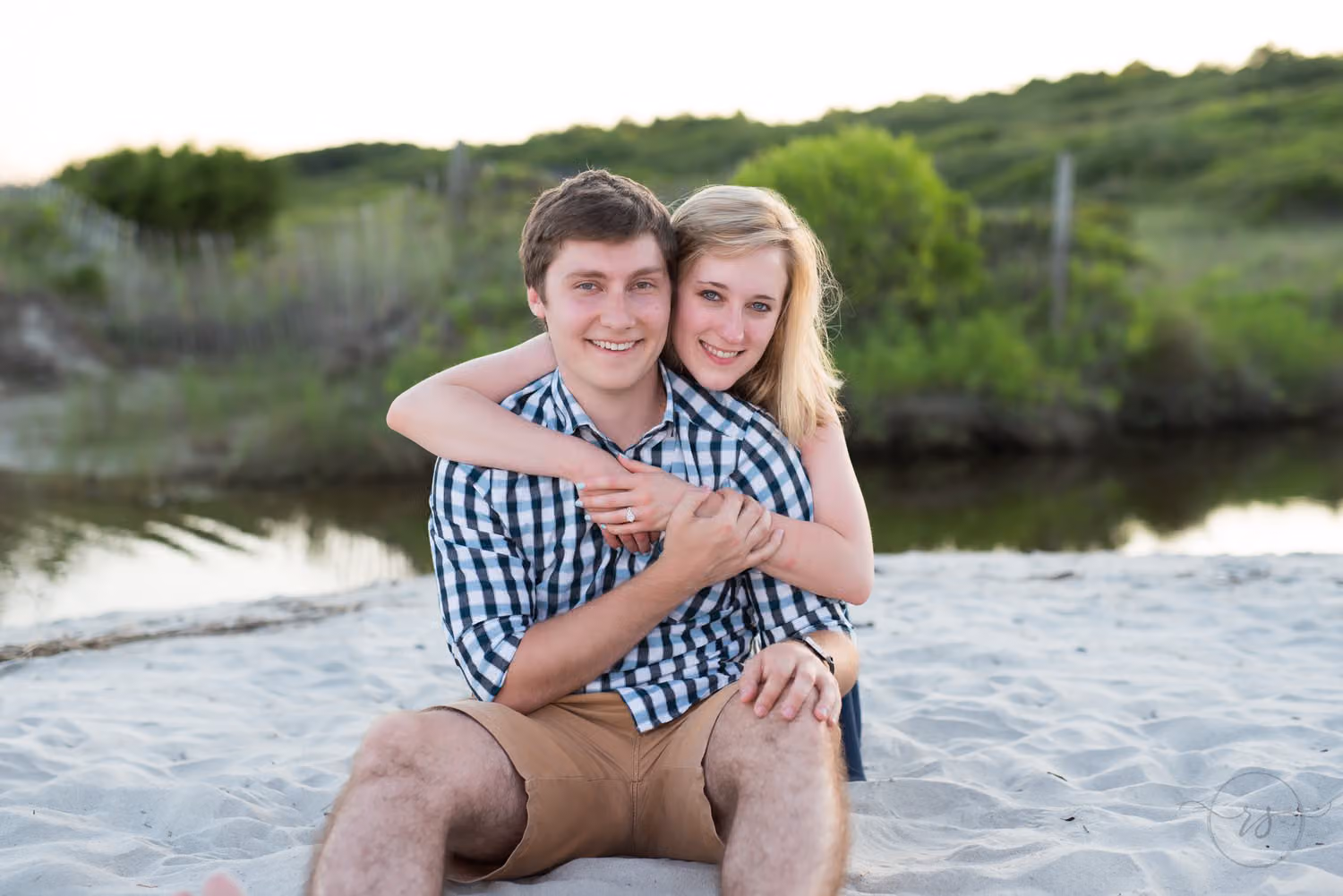 Newly engaged woman hugging her fiancé from behind as they sit in the sand by a tidal pool at Myrtle Beach State Park