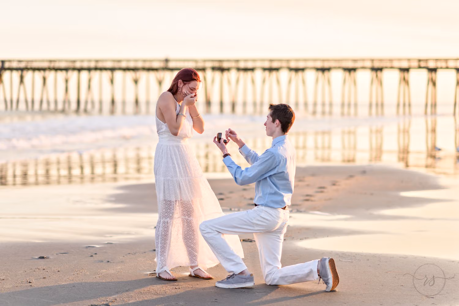 Man down on one knee holding out a ring as she covers her face, with the pier behind them at Myrtle Beach State Park