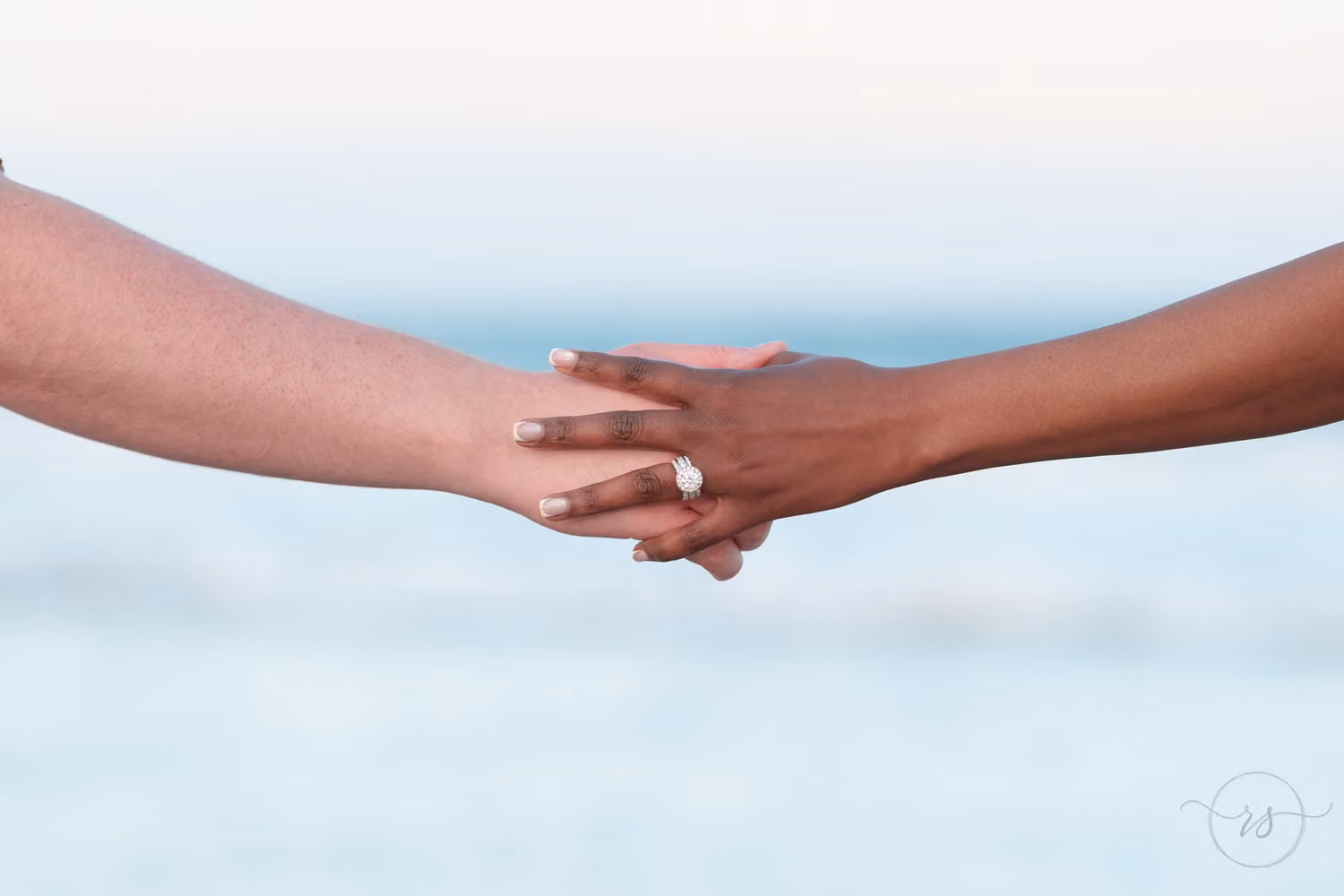 Close view of two hands clasped to show off an engagement ring against the blue ocean in Pawleys Island
