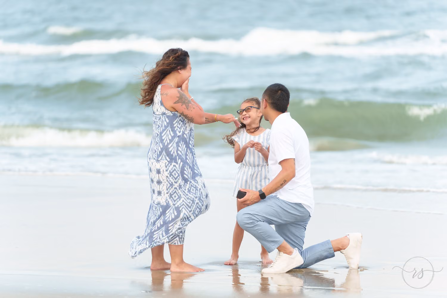 Woman reaching for her little girl while her fiancé kneels in the surf at Huntington Beach State Park