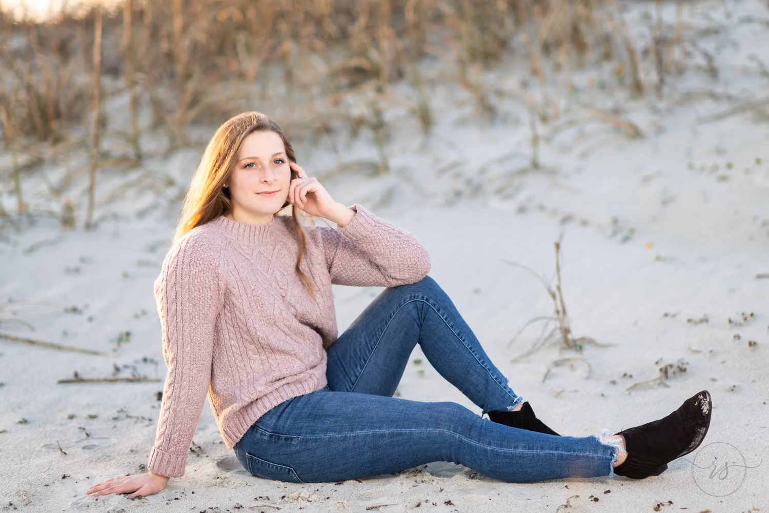Senior in a pink sweater sitting in the sand by the dunes with her hand at her chin at Huntington Beach State Park