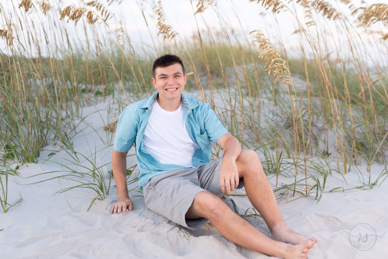 Senior guy in a teal shirt and shorts sitting barefoot in the sand by the sea oats at Huntington Beach State Park