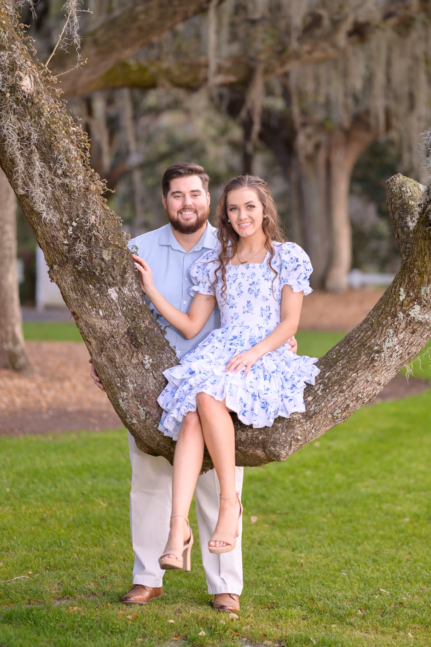 Woman in a blue floral dress sitting on a low oak branch beside her standing fiancé at Caledonia Golf and Fish Club