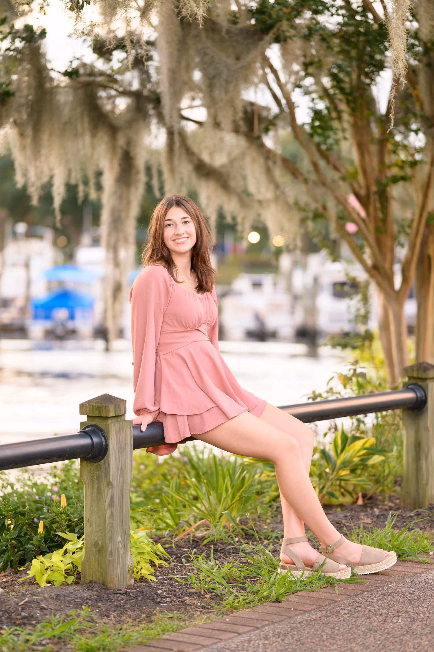 Senior girl in a pink dress sitting on a railing by the river under moss-draped oaks at the Conway River Walk