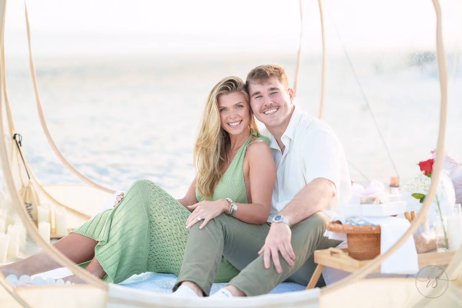 Couple sitting close on cushions inside a clear dome tent with a picnic spread at Huntington Beach State Park
