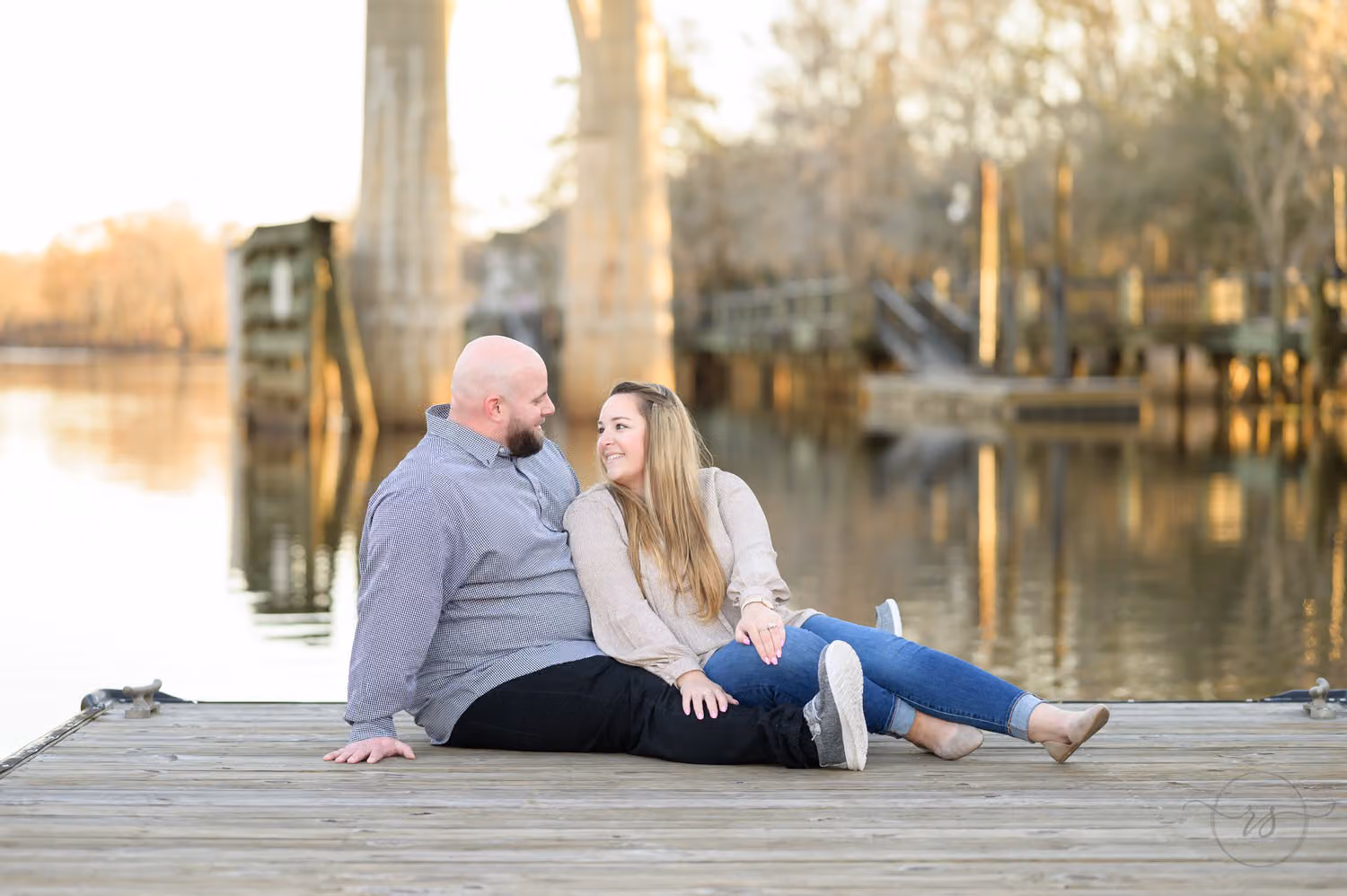 Couple sitting close together on a wooden dock with the river and old pilings behind them along the Conway Riverwalk