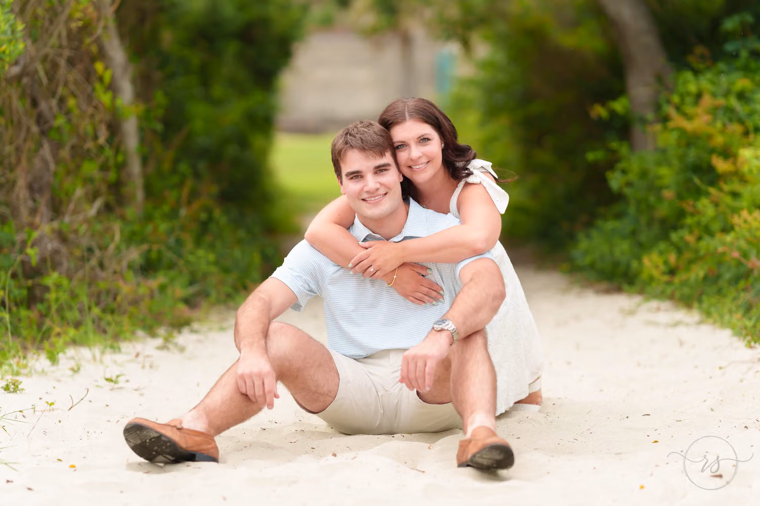 Woman hugging her fiancé from behind as they sit smiling on a sandy path under the trees at Huntington Beach State Park