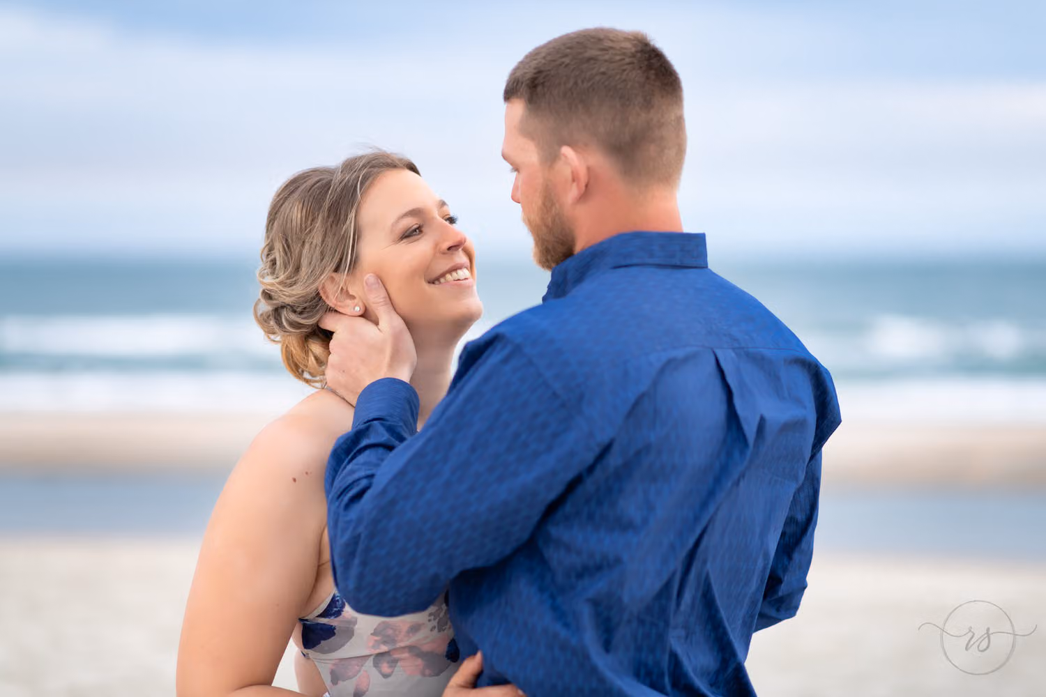 Woman smiling up at her fiancé in a blue shirt as he touches her face by the ocean at Huntington Beach State Park