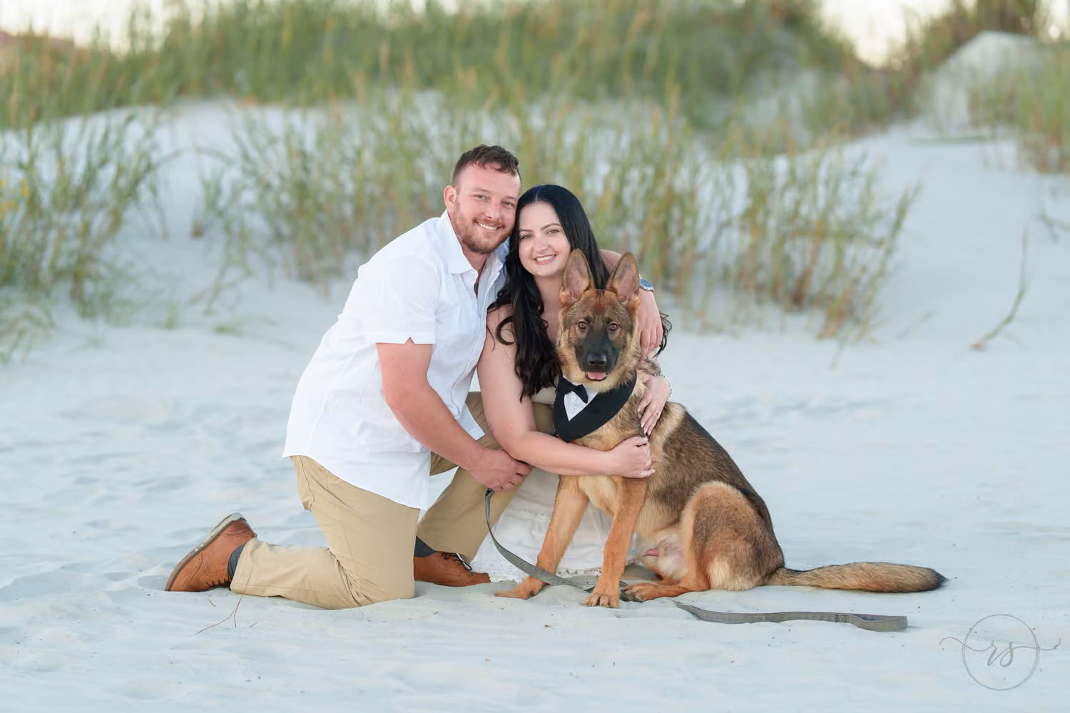 Smiling couple kneeling in the sand by the dunes, hugging their German shepherd wearing a bow tie collar